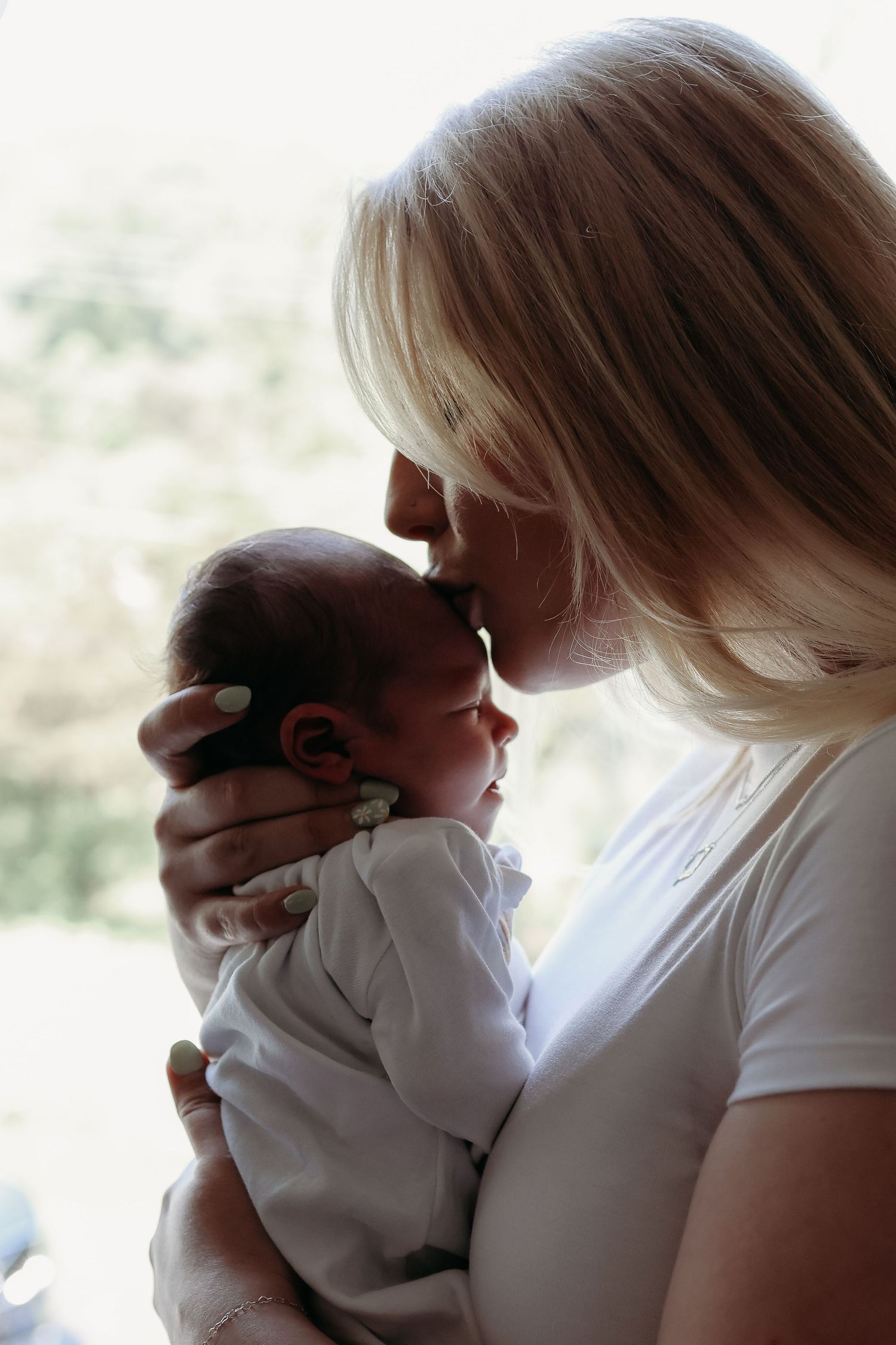 Blonde mother tenderly kissing her newborn baby near a bright window in soft light.