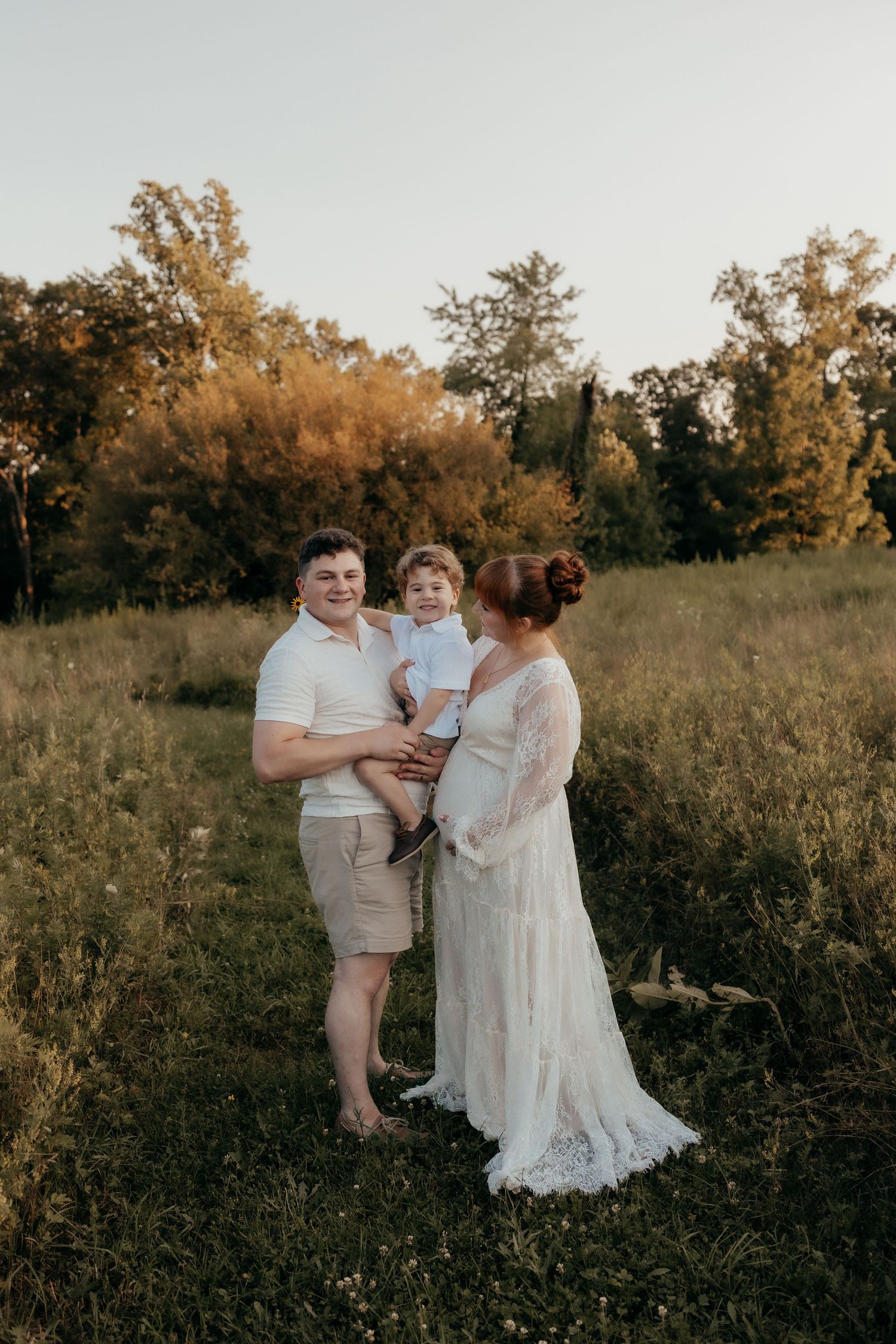 A family of three poses in a golden field at sunset, with a man holding a toddler beside a pregnant woman in lace gown.