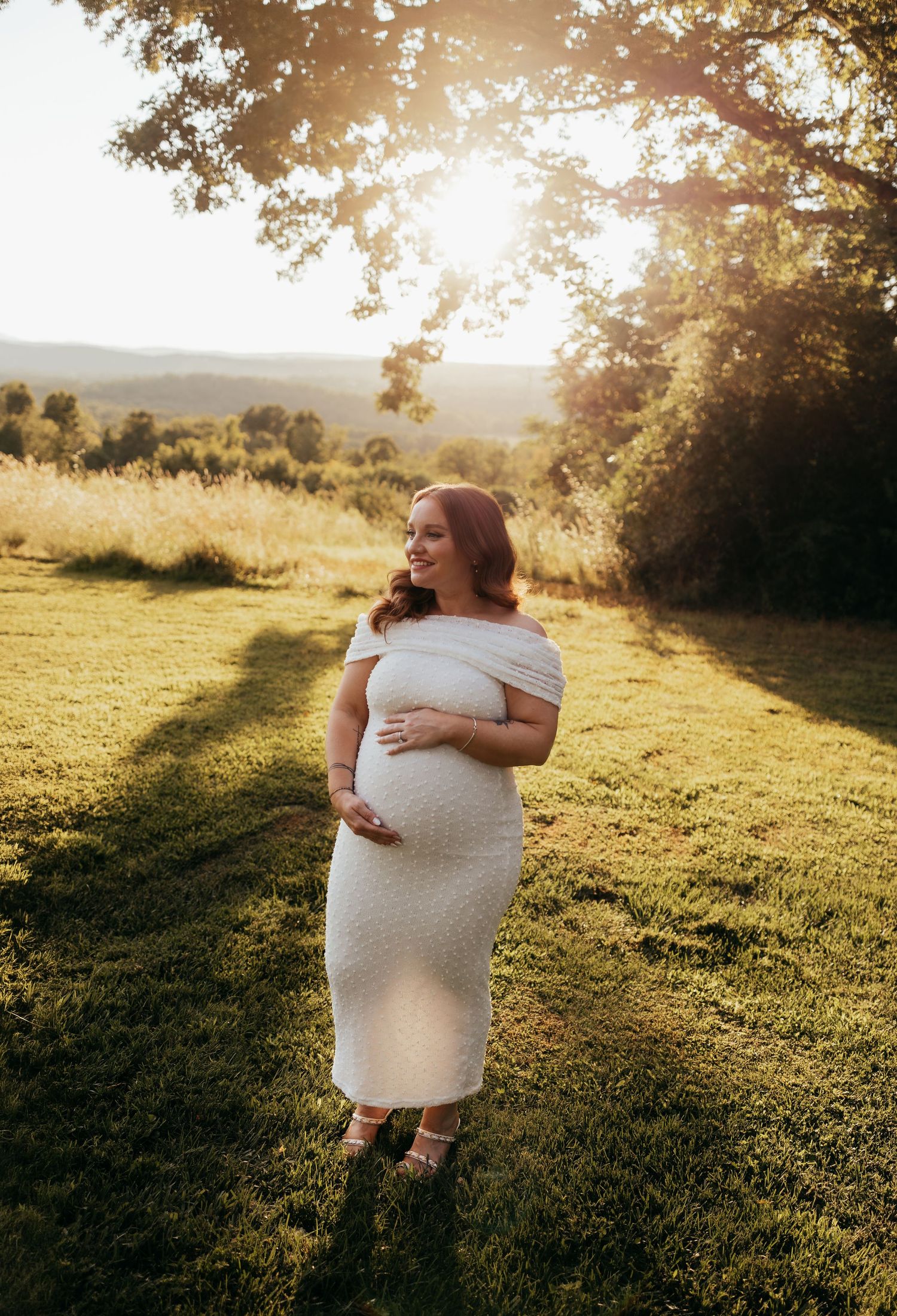 Pregnant woman in white off-shoulder dress glowing in golden sunset light on lush green hillside.