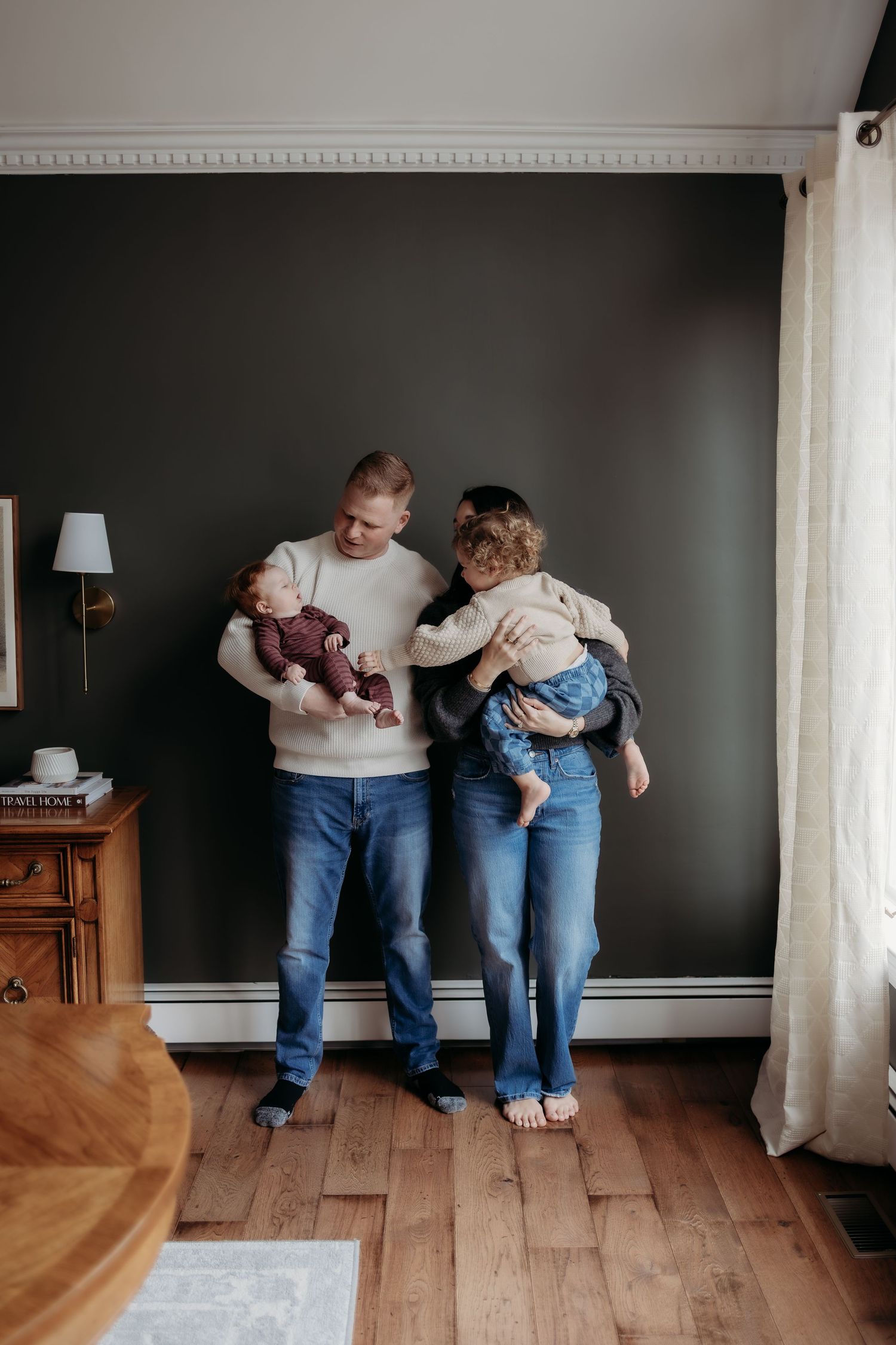 A young family of four poses indoors, parents holding two small children against a dark wall with hardwood floors.