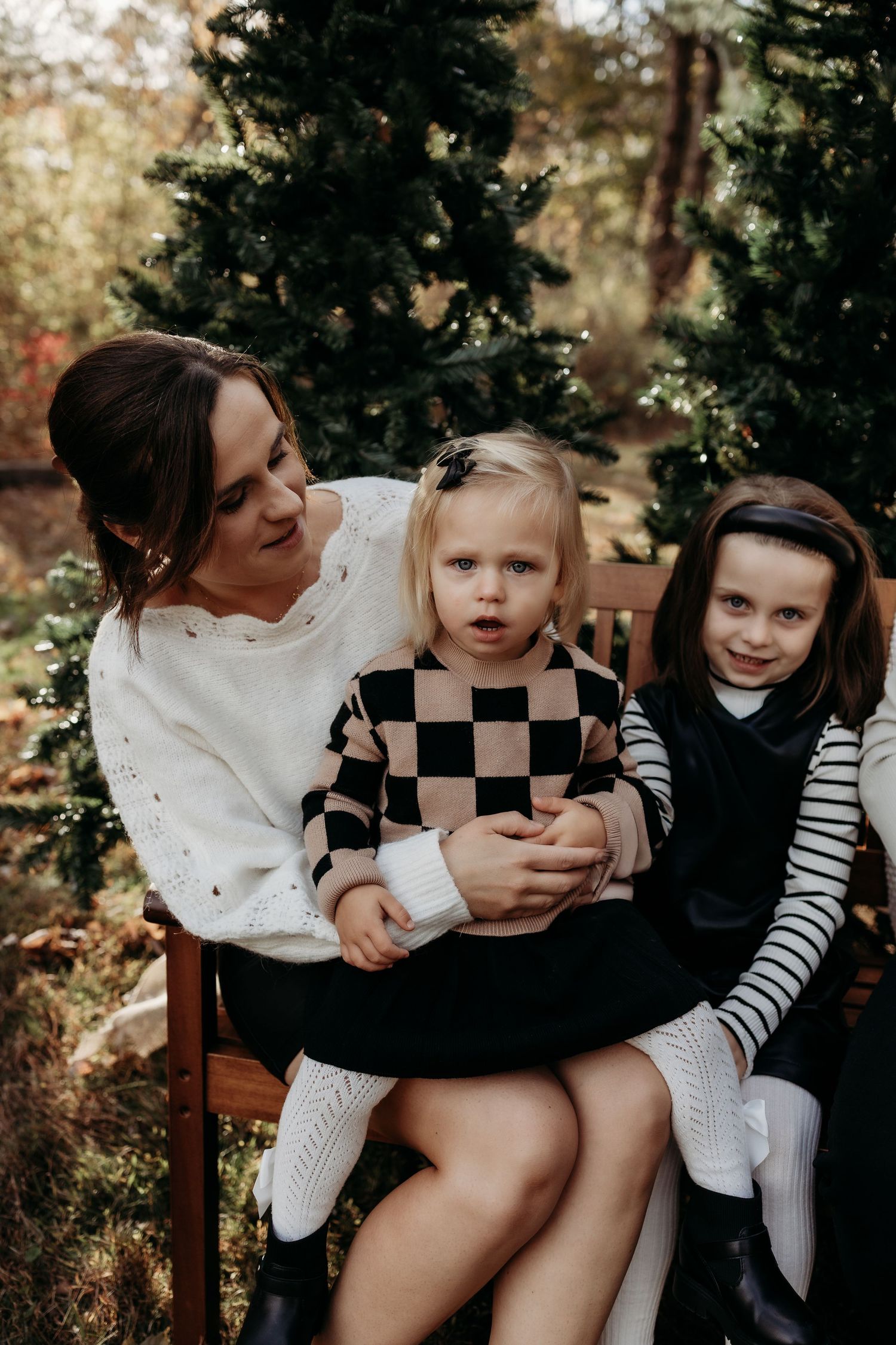 A mother sits on a bench holding two young daughters near evergreen trees in an outdoor holiday family portrait.