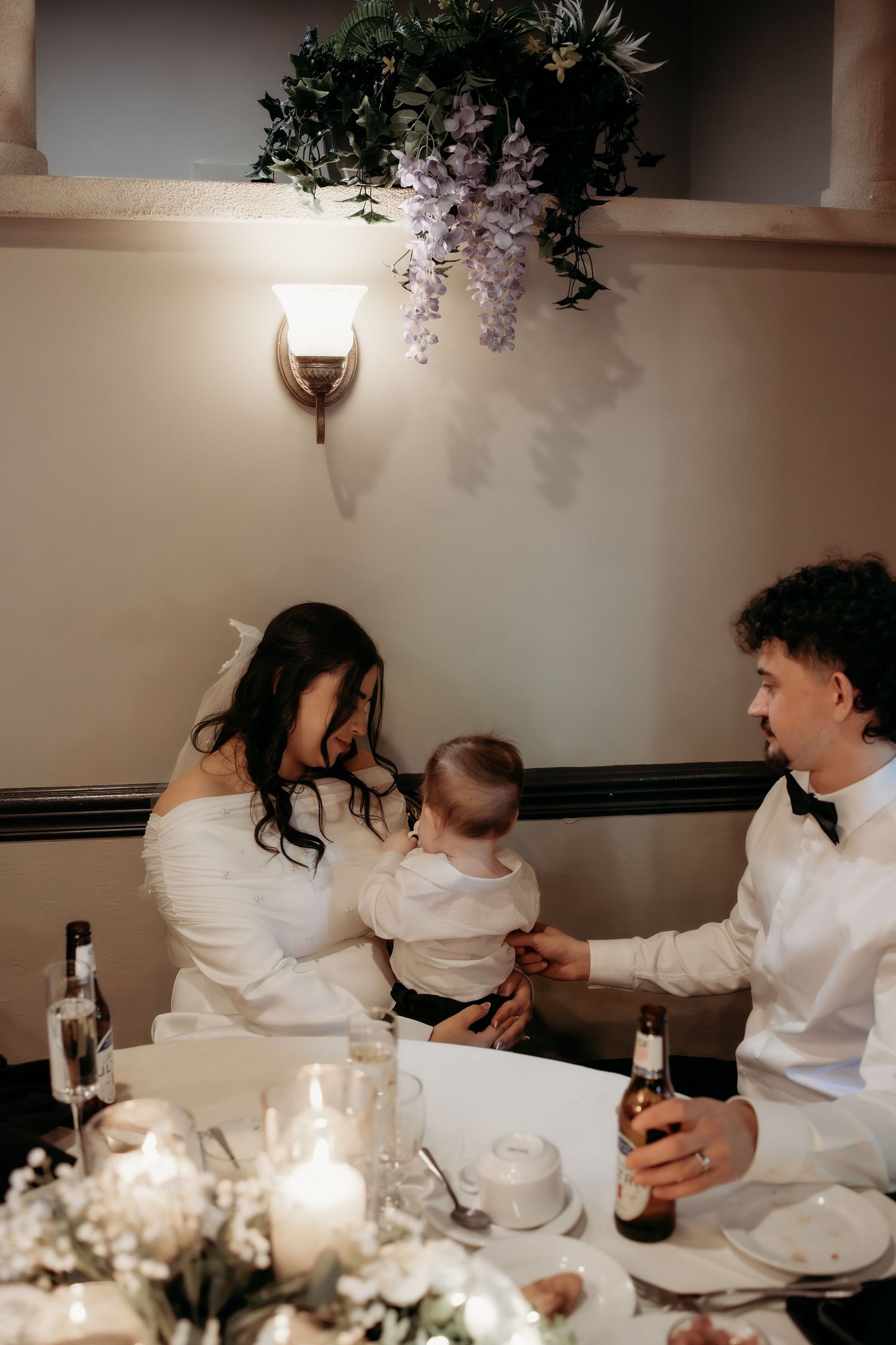 Newlywed couple with baby at candlelit wedding reception table adorned with floral arrangements.