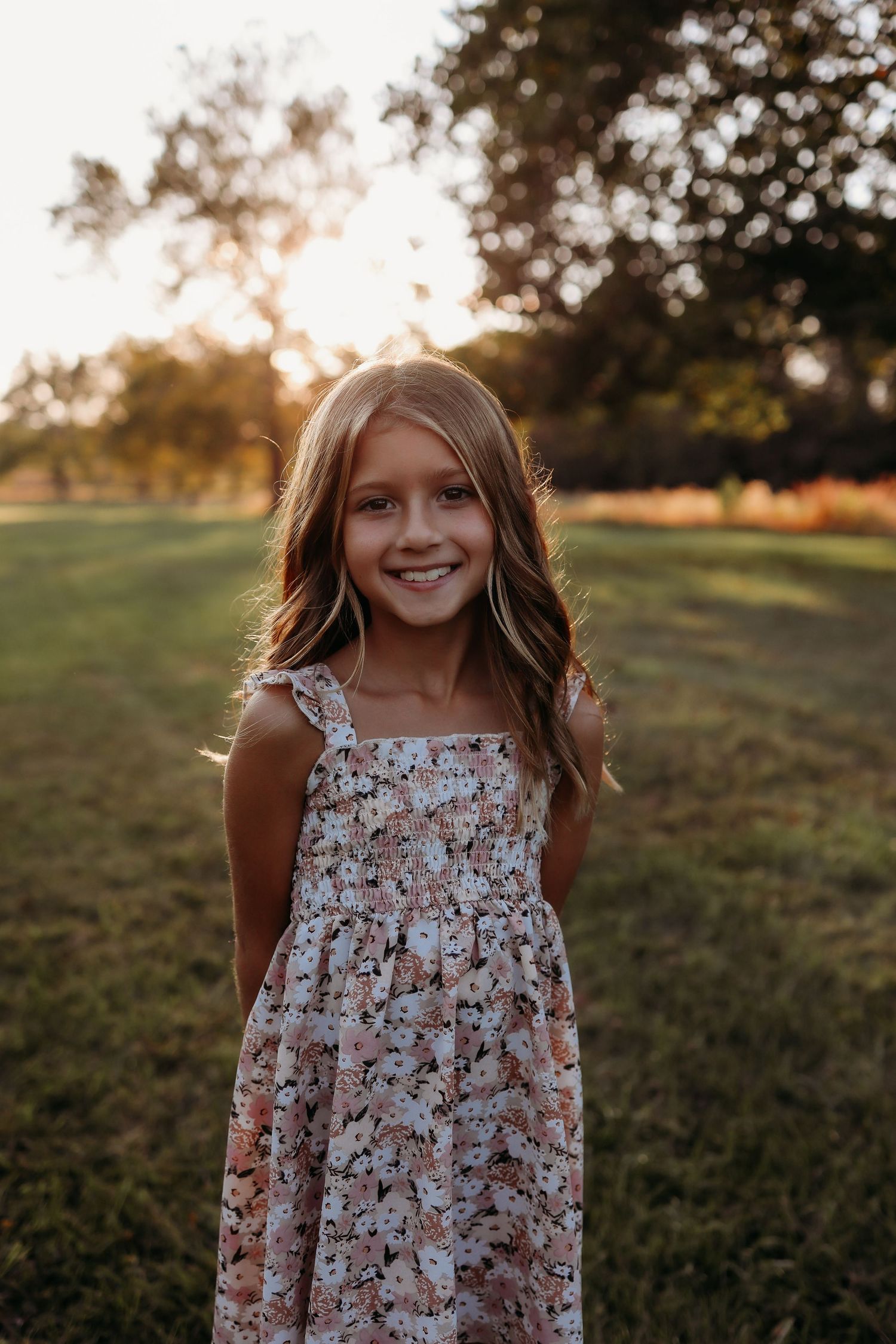 Smiling young girl in floral sundress standing in green park during golden sunset.