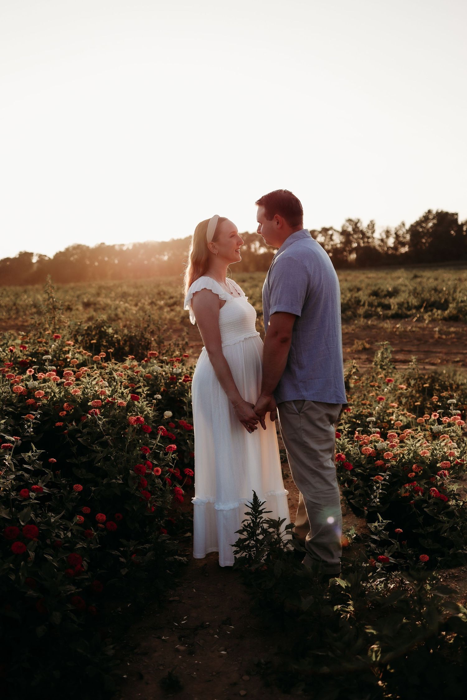 Expecting couple holds hands and shares a tender moment in a vibrant flower field at golden sunset.
