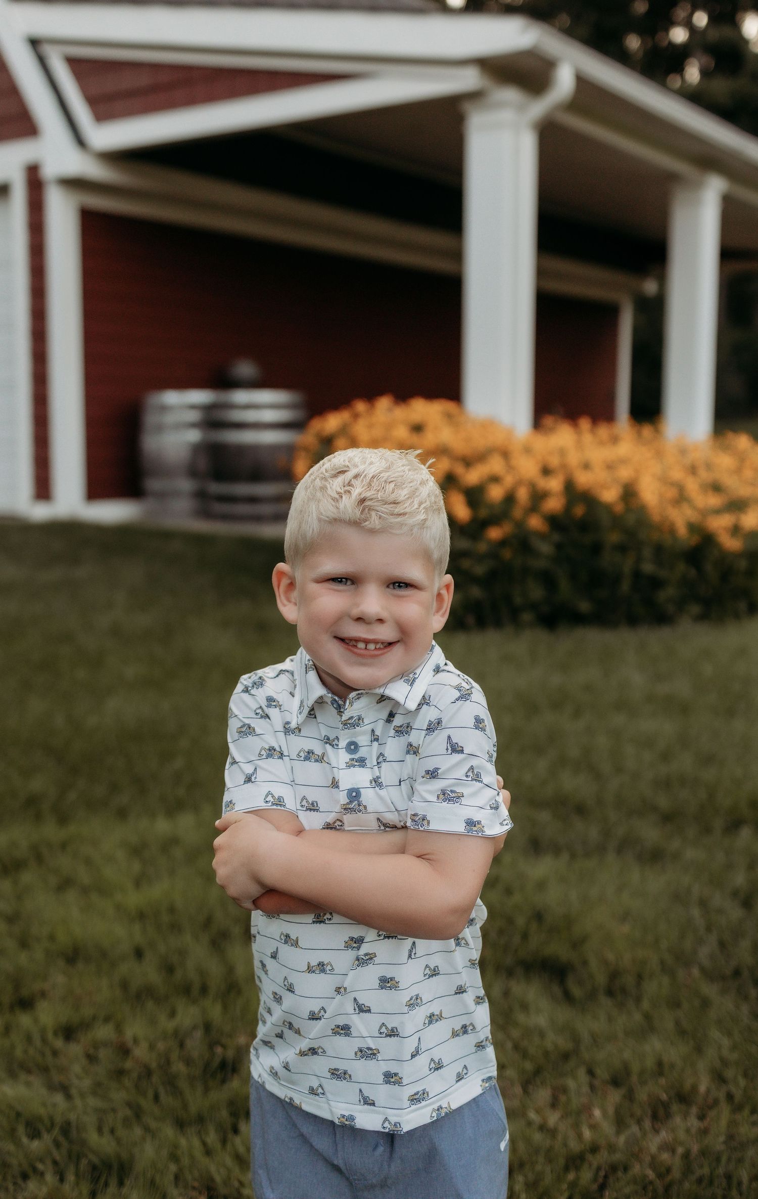Smiling blonde boy in printed shirt stands arms crossed on green lawn near red barn with orange flowers.