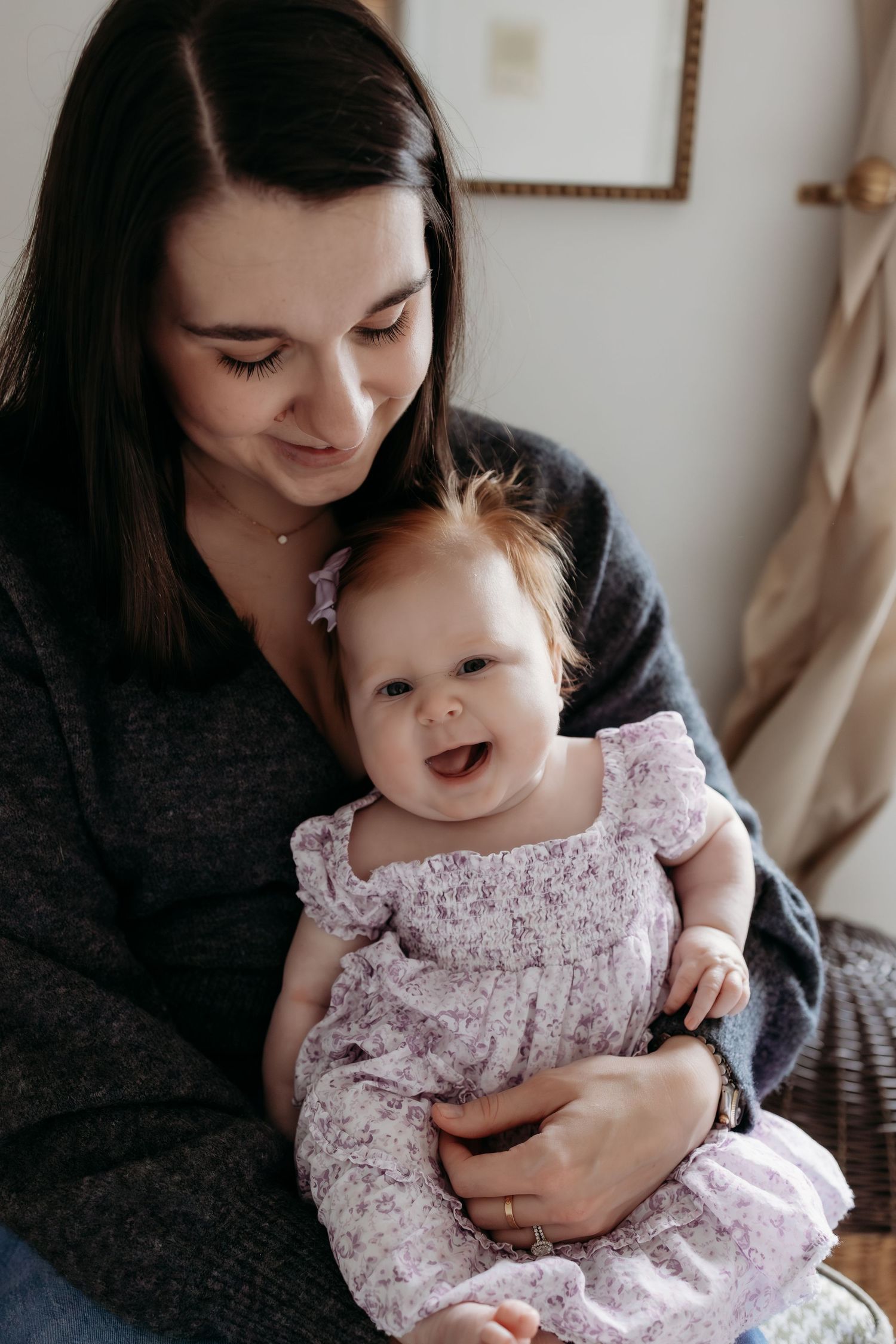 Smiling baby girl in floral dress being held by her dark-haired mother indoors.
