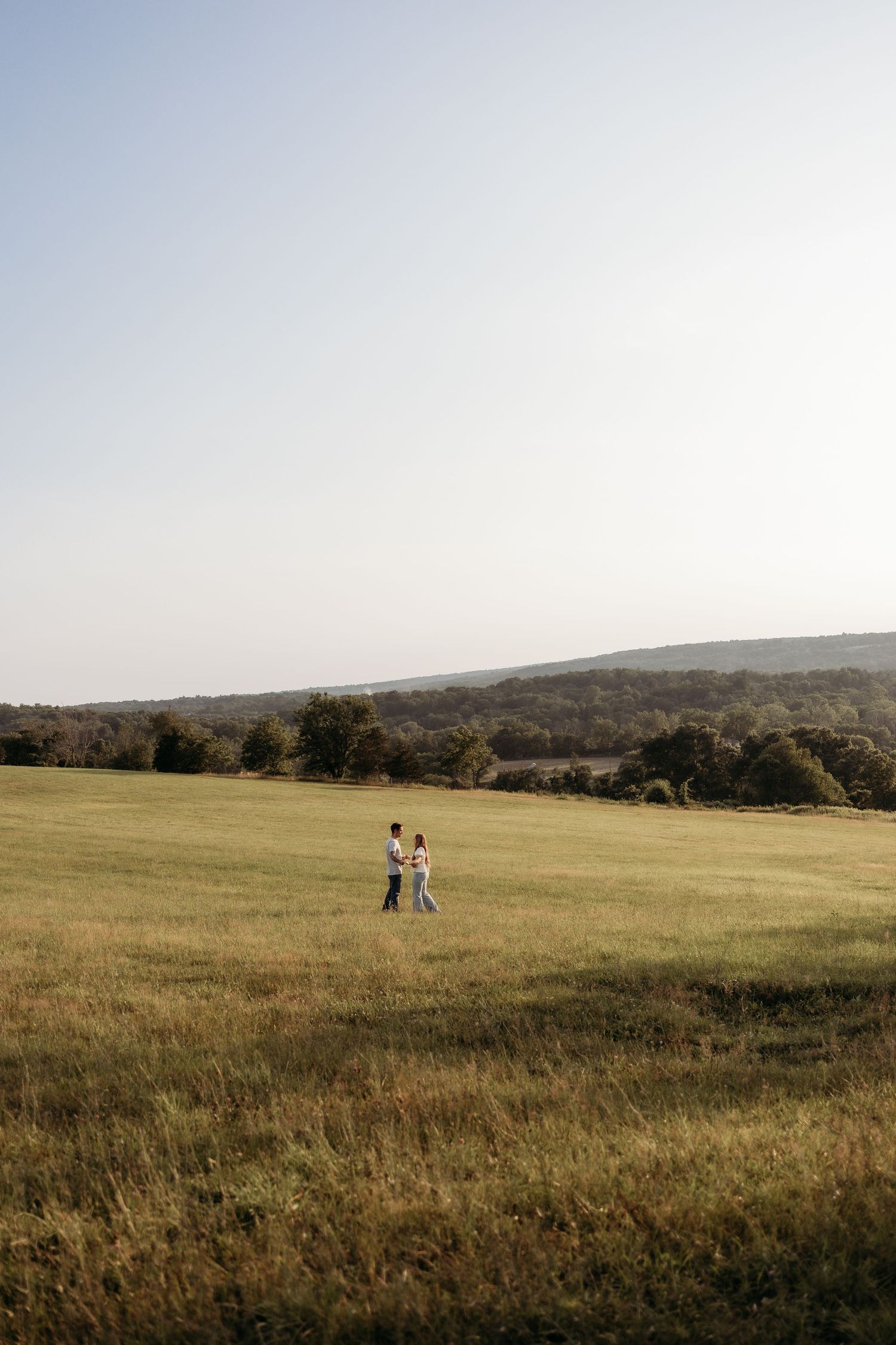 Couple walking hand in hand through a sunlit green meadow with rolling hills and trees in the background.
