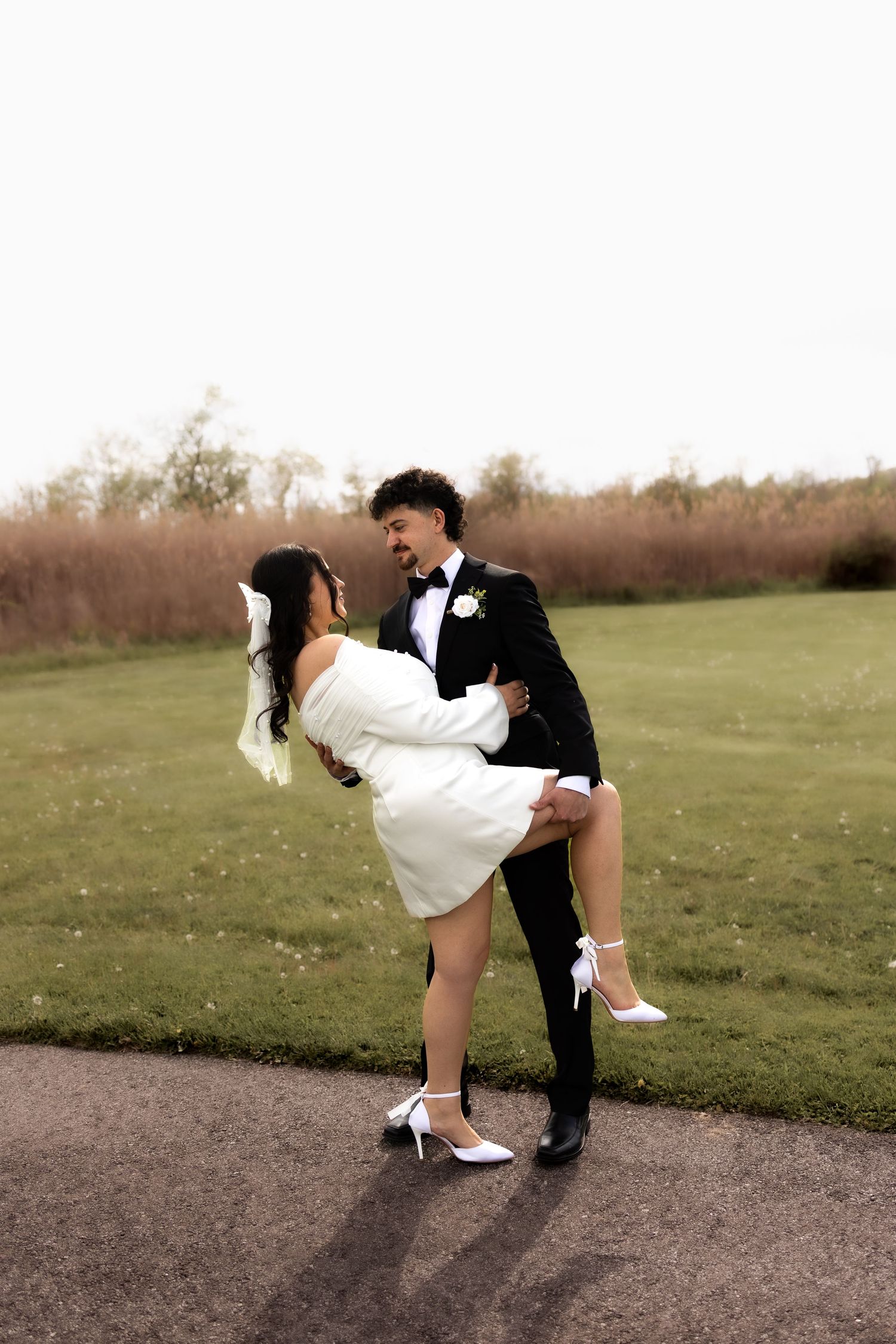 Groom lifting smiling bride in white mini dress outdoors on their wedding day.