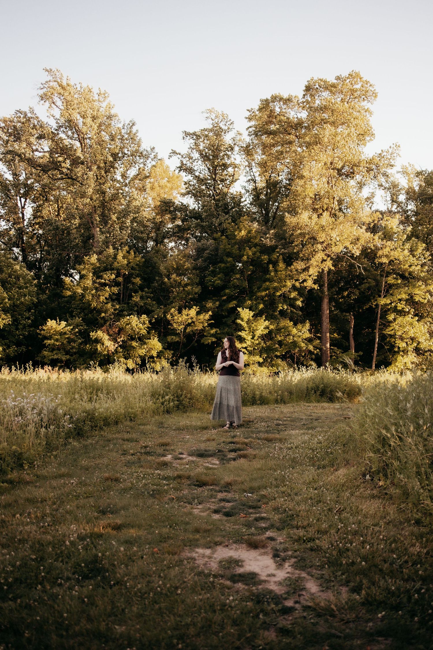 Woman in dark top and maxi skirt stands on a dirt path in a lush green meadow surrounded by tall trees.