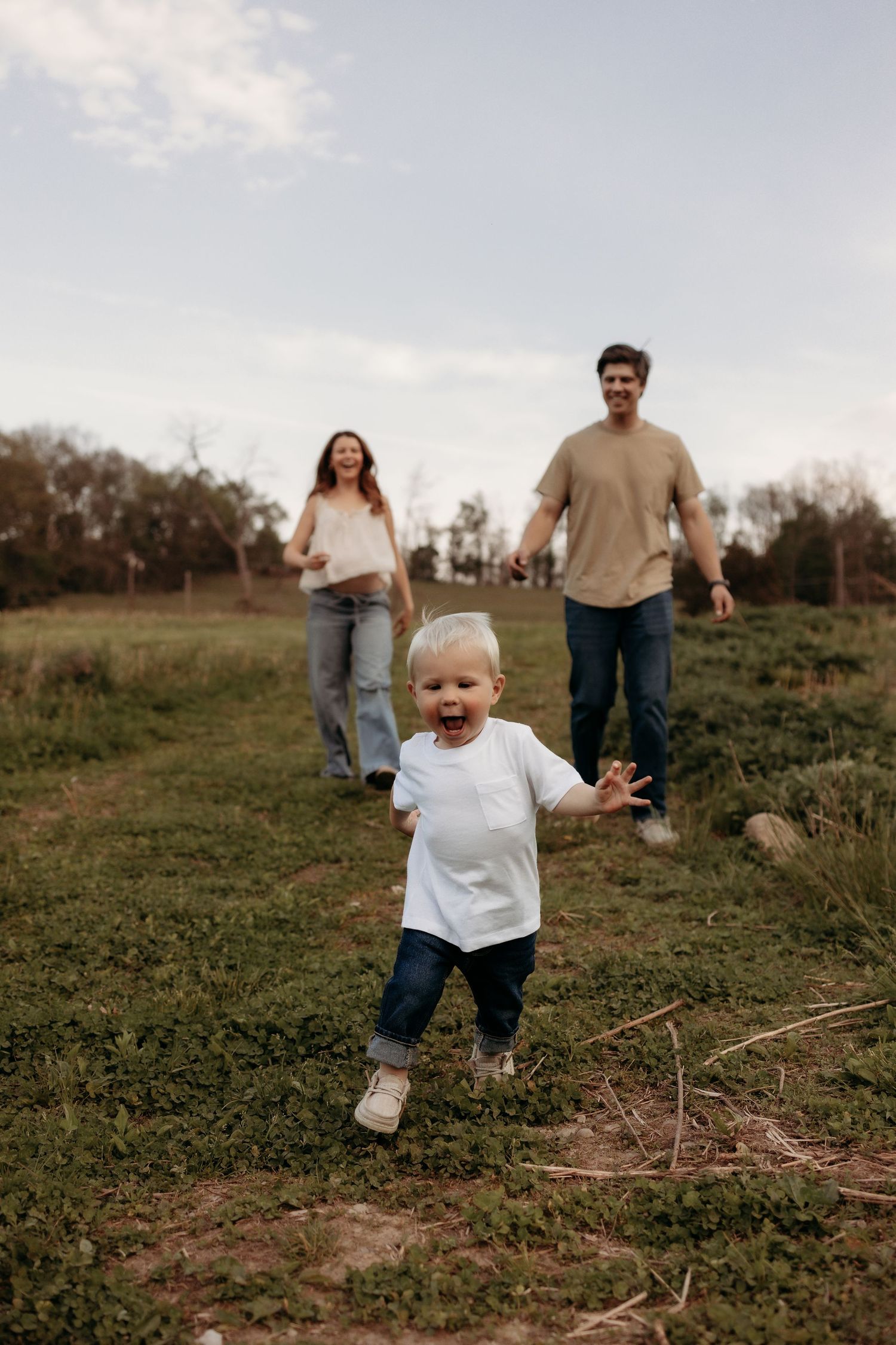 Young family with excited toddler running in green field outdoors.