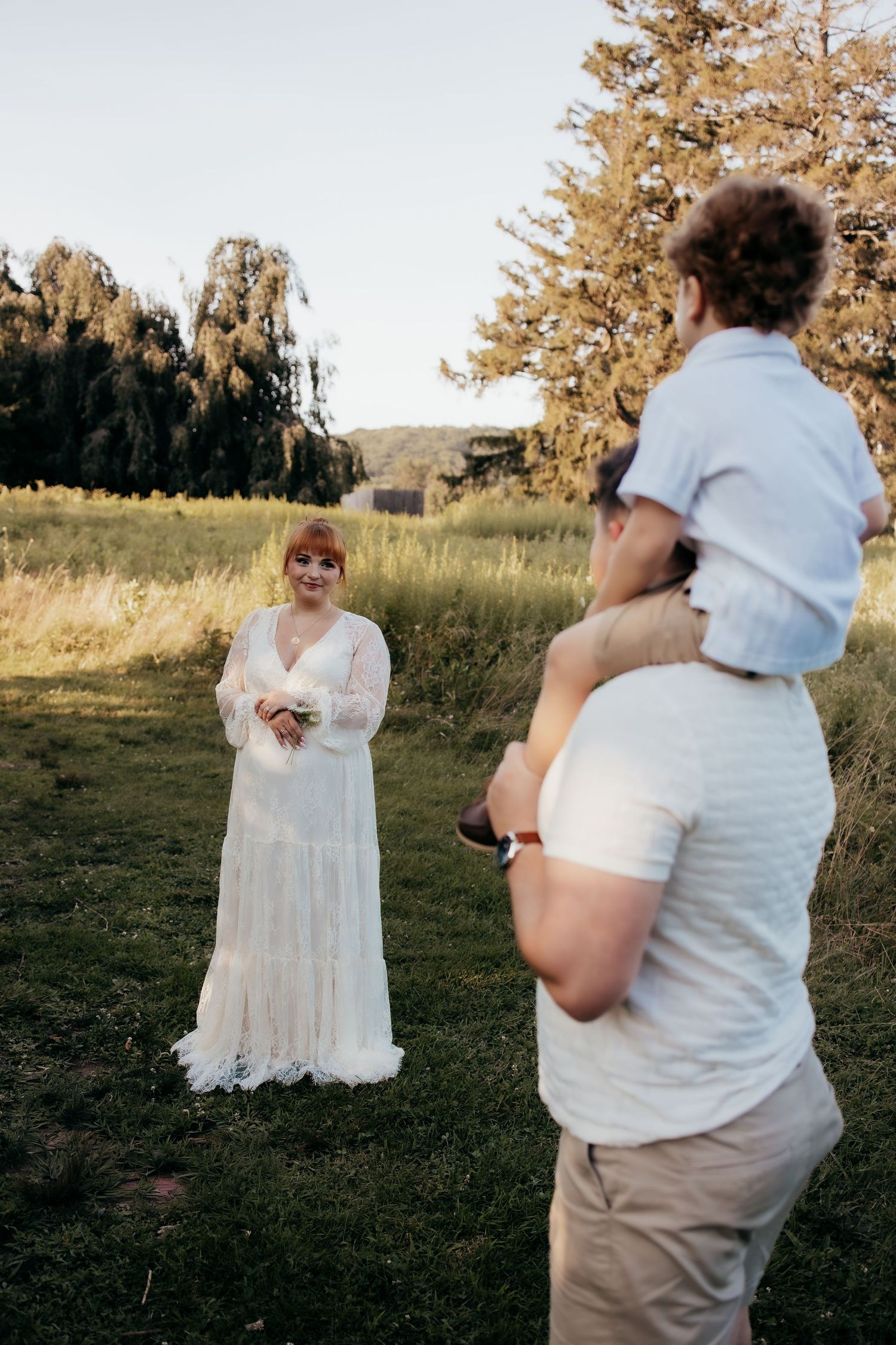 Bride in white lace gown smiles at man holding child on shoulders in a green field at golden hour.