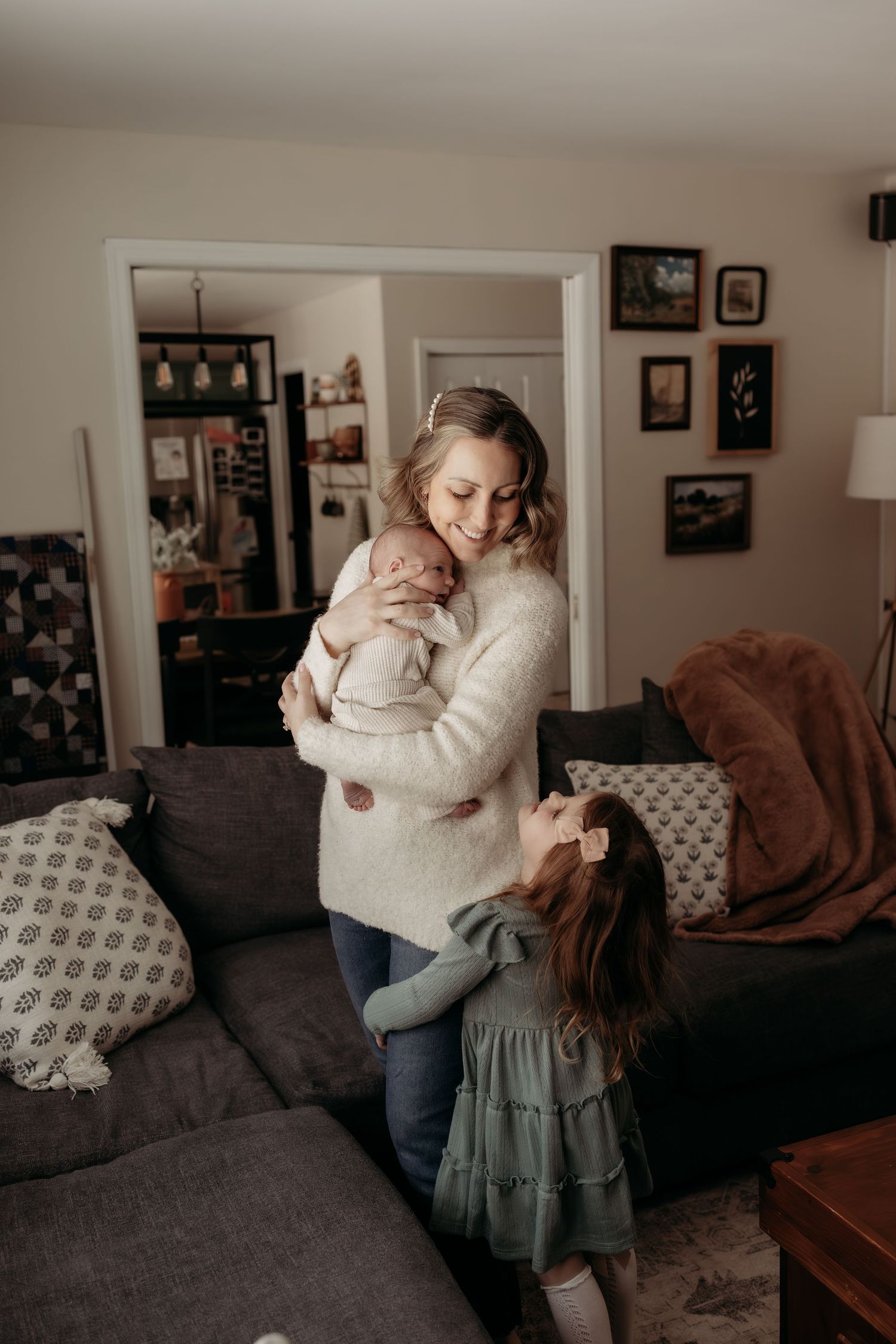 Smiling mother in cream sweater holds newborn while toddler daughter hugs her in living room.