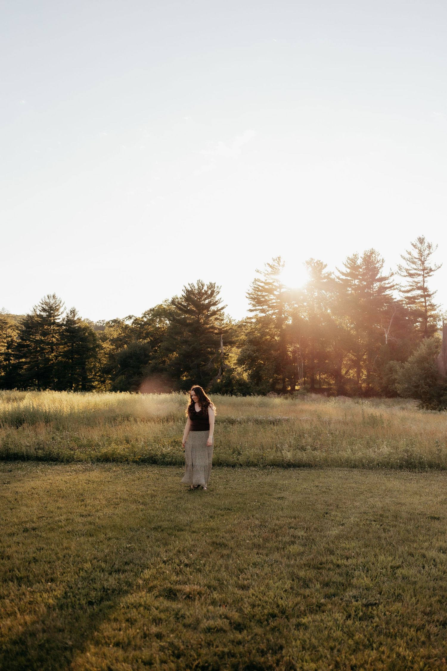 Woman in black top and maxi skirt stands alone in open field with pine trees and golden sunset behind her.