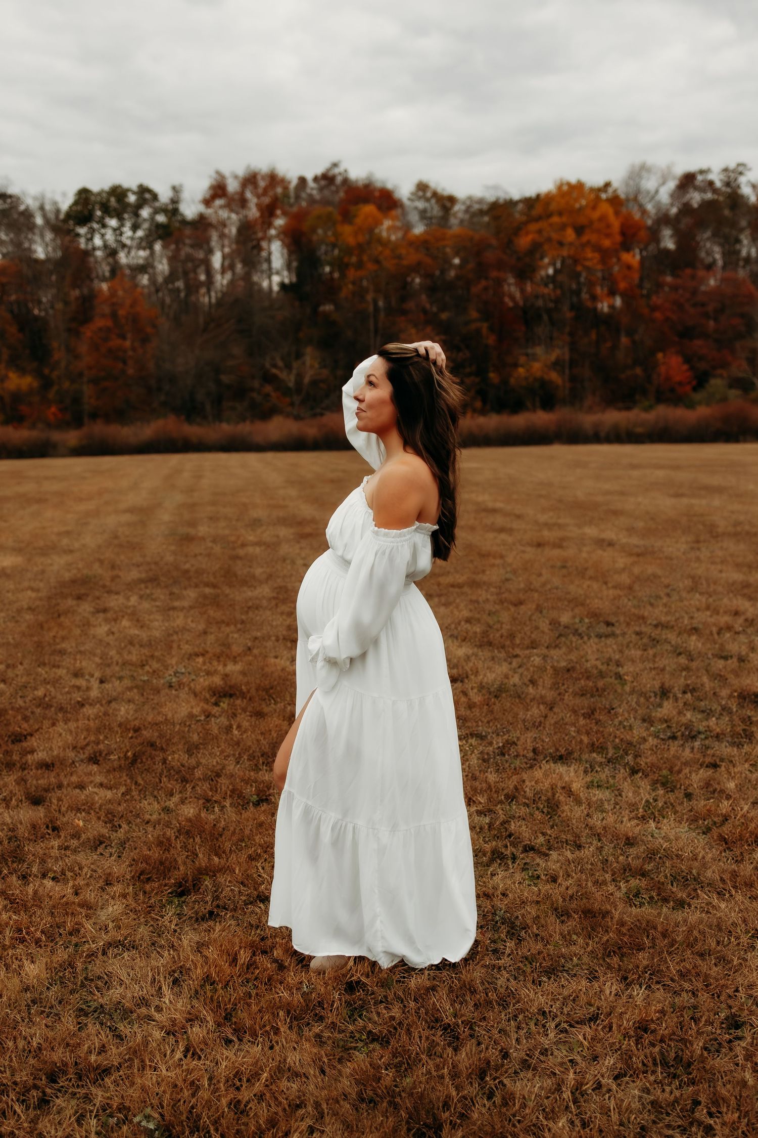 A woman in a flowing white off-shoulder dress stands in an autumn field with orange trees in the background.