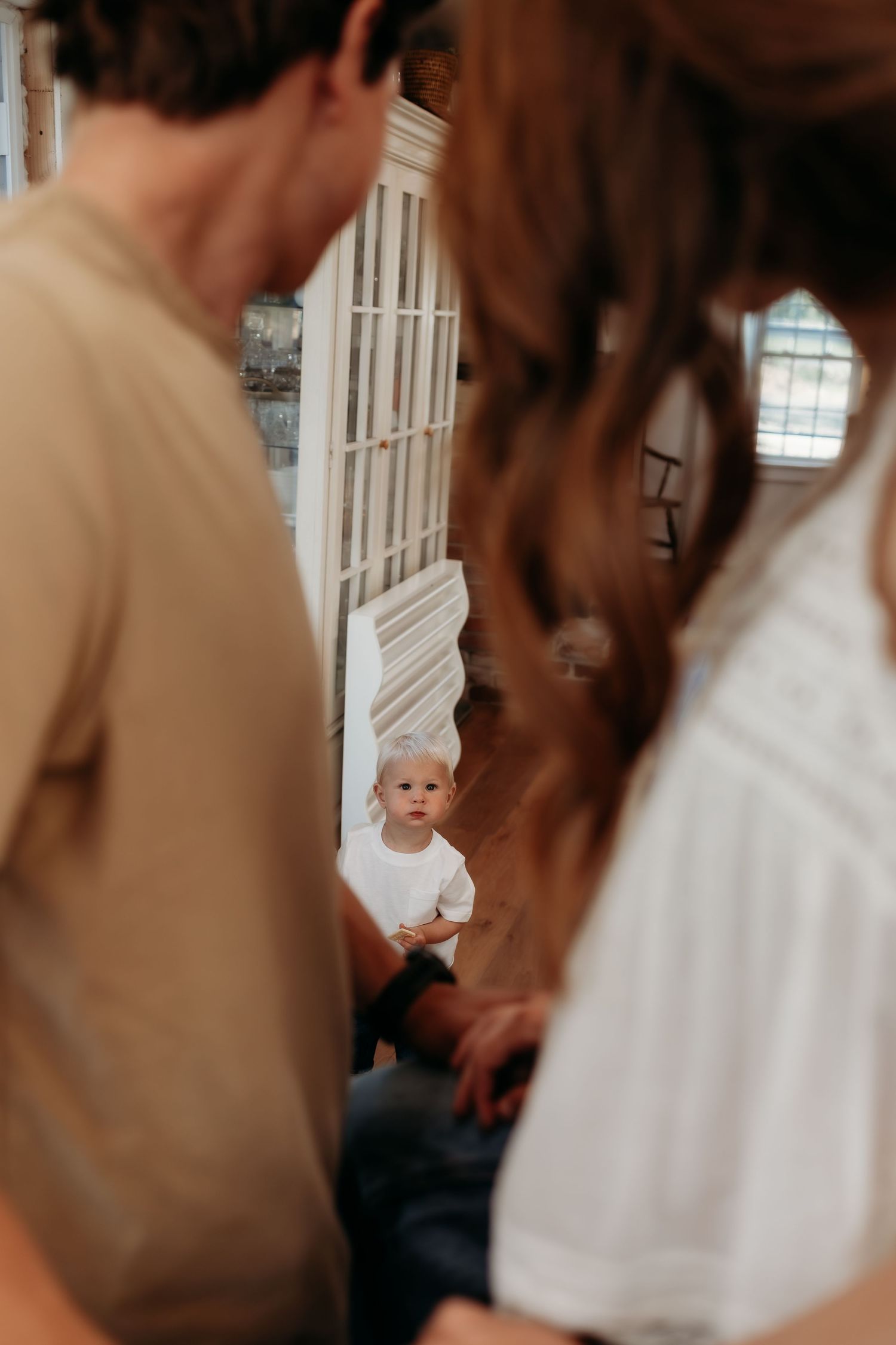 A toddler in a white shirt stands between two adults in a warmly lit home interior.