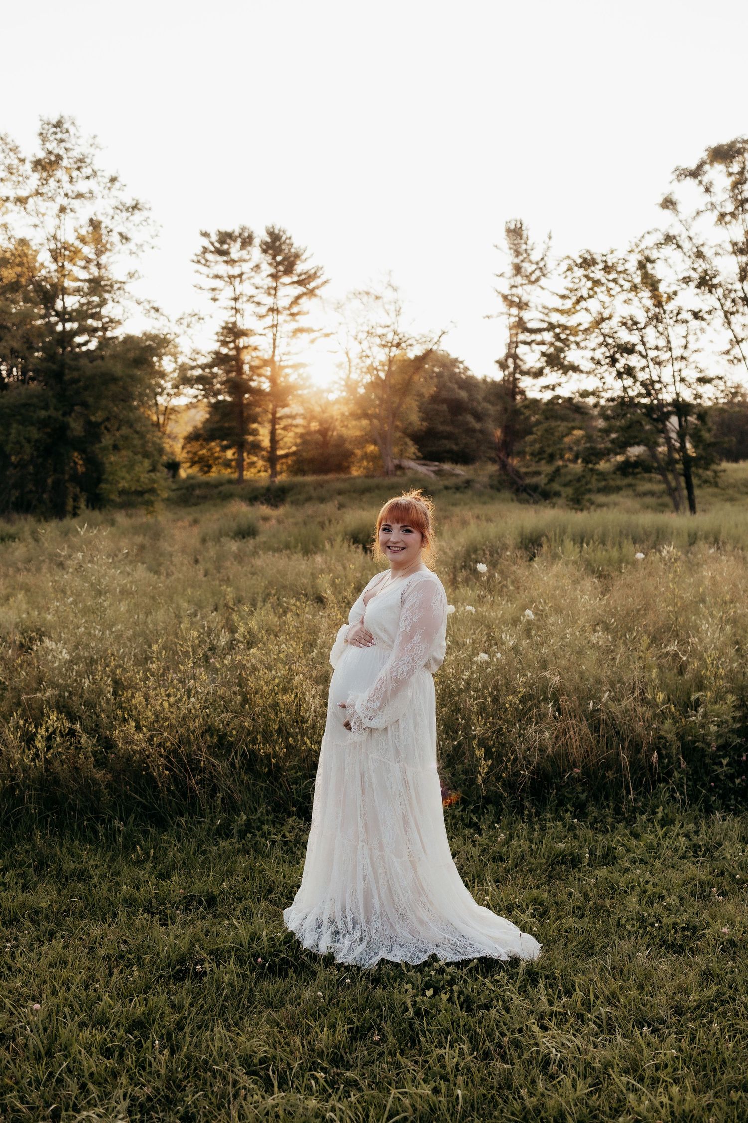 A pregnant woman in a flowing white lace maternity gown poses in a sunlit field at golden hour.