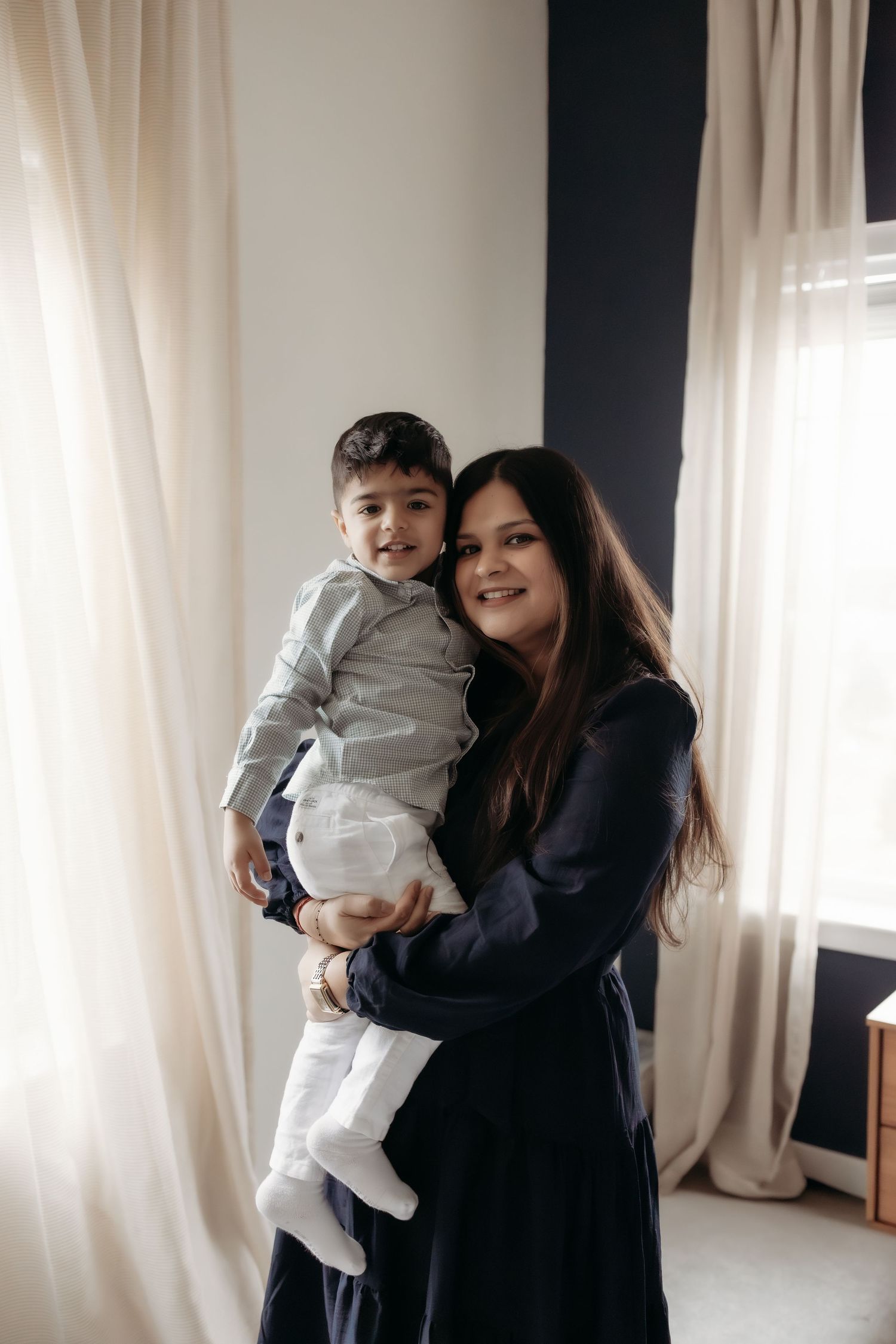 Smiling mother in navy dress holds happy toddler boy in gray shirt near bright window.