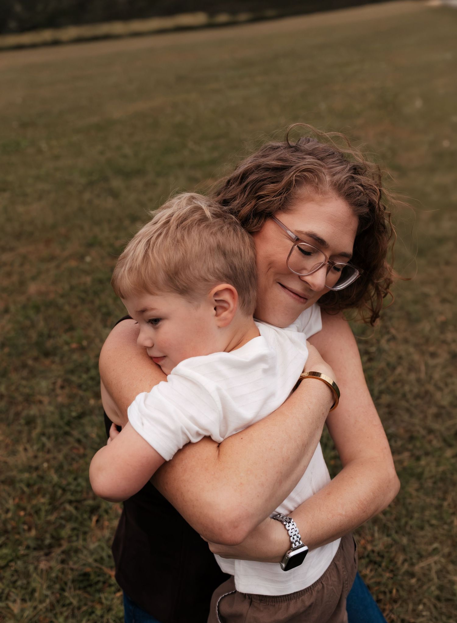 A smiling mother with curly hair and glasses warmly hugging her young blonde son in an open grassy field.