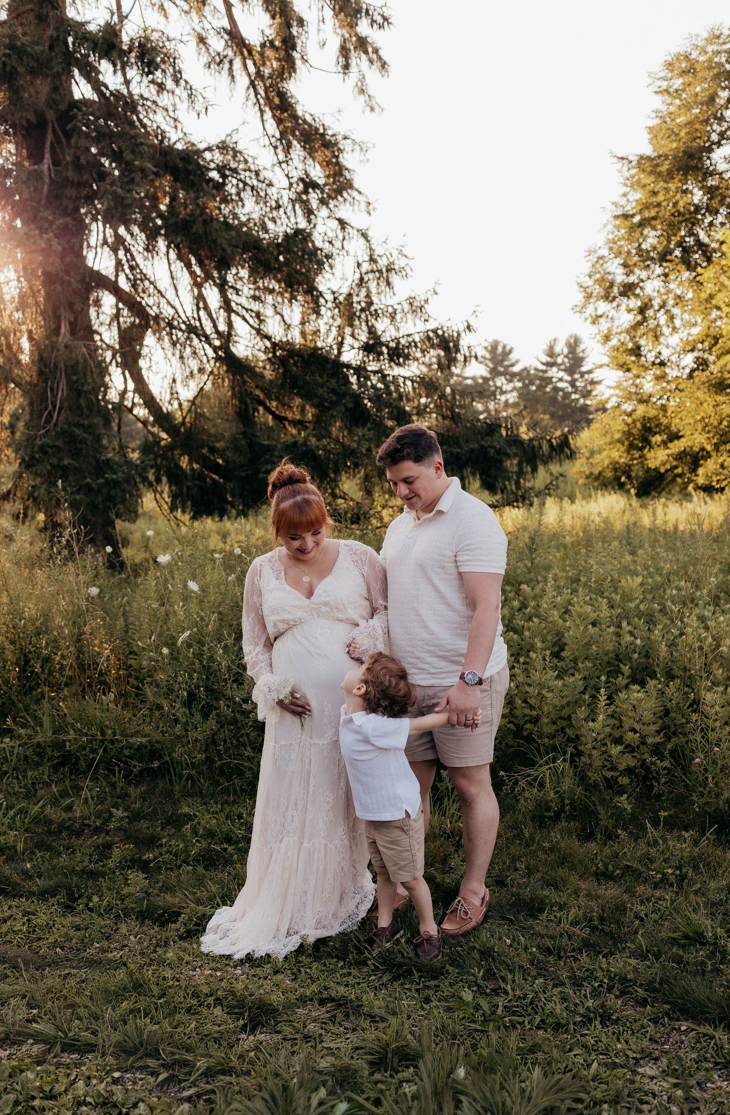Pregnant woman in white dress with husband and toddler during golden hour maternity photoshoot in lush green field.