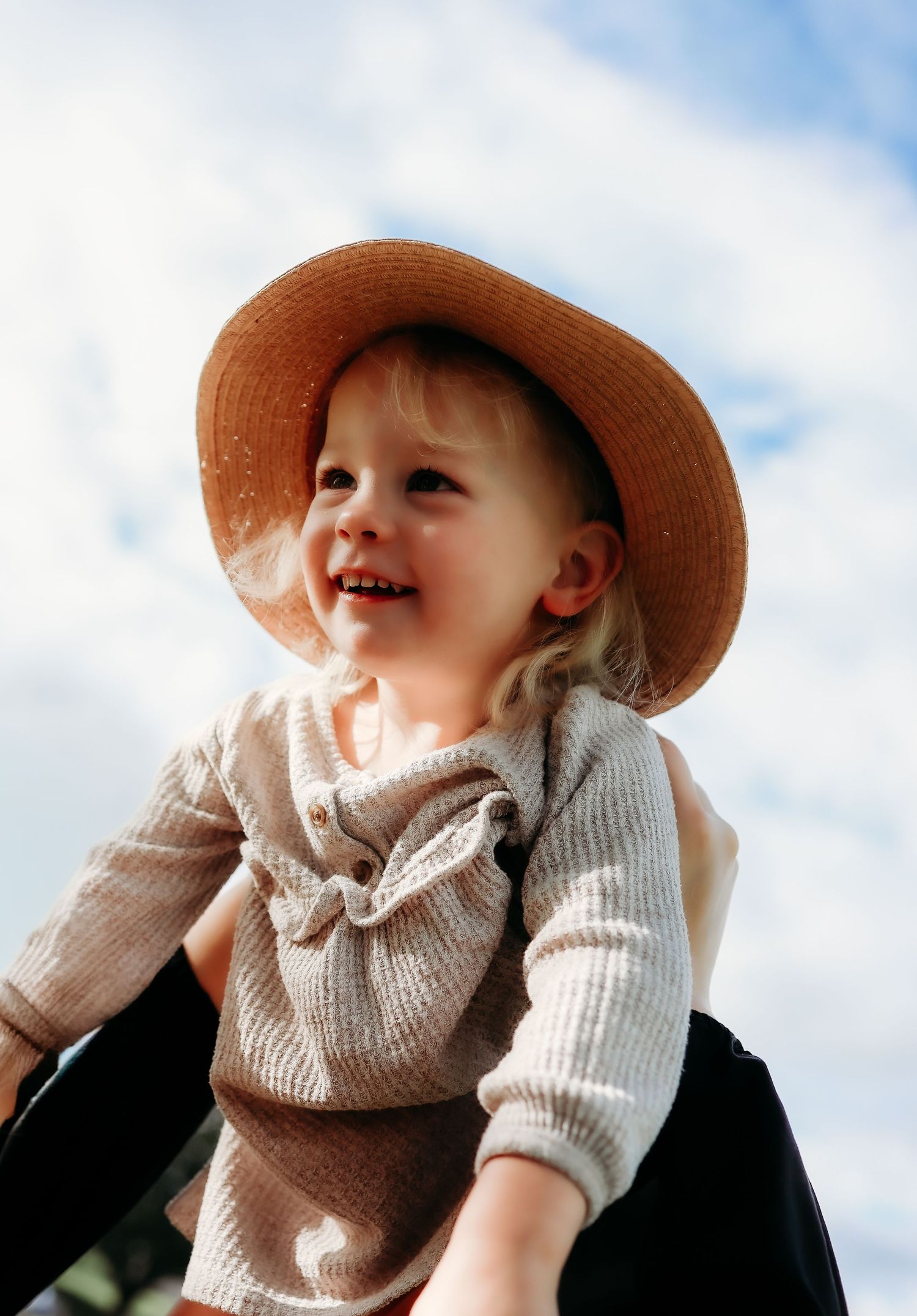 Joyful toddler girl in a straw hat laughs while being lifted against a bright blue sky.
