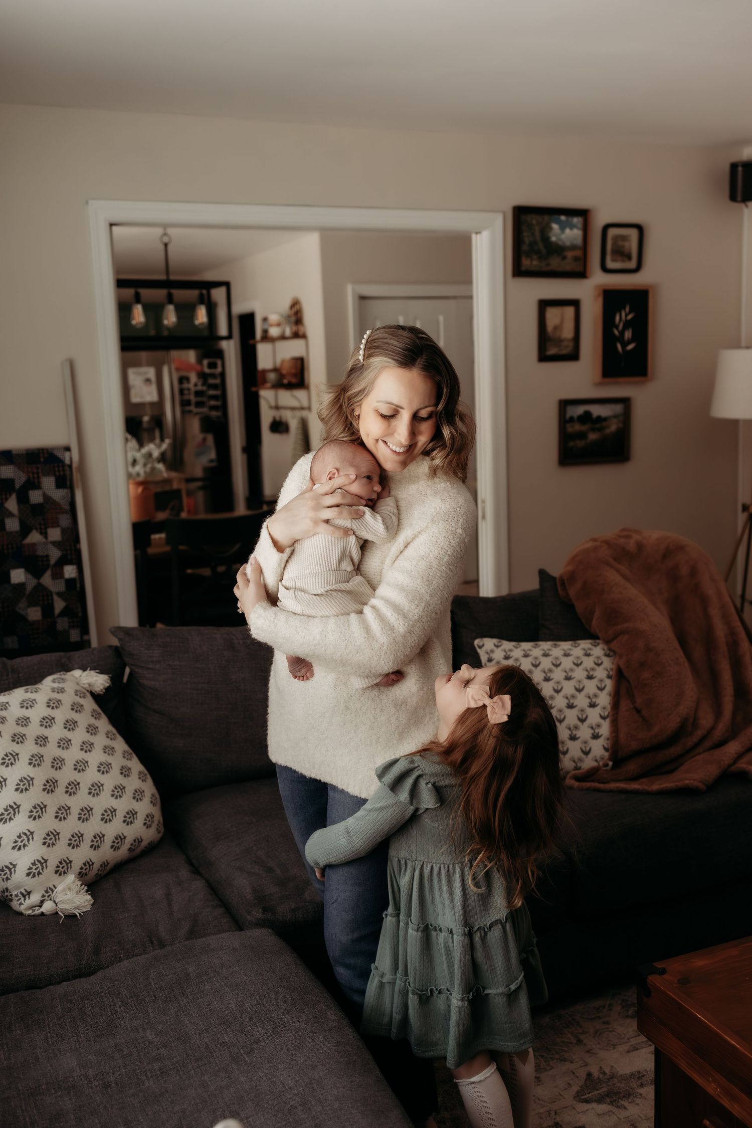 Blonde mother in white sweater holding newborn while young daughter hugs her in living room.