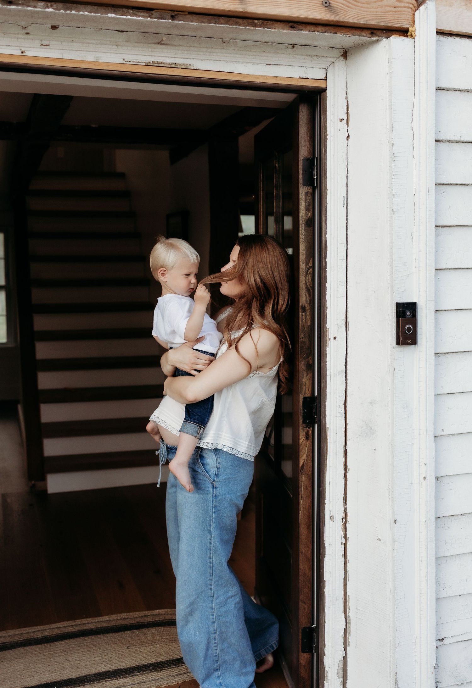 A woman in jeans holds a toddler in a doorway of a white farmhouse, sharing a tender moment.