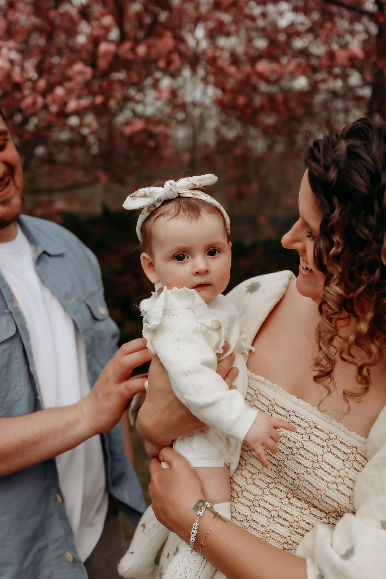Parents admiring baby girl in white outfit with bow headband near pink blossoming trees.