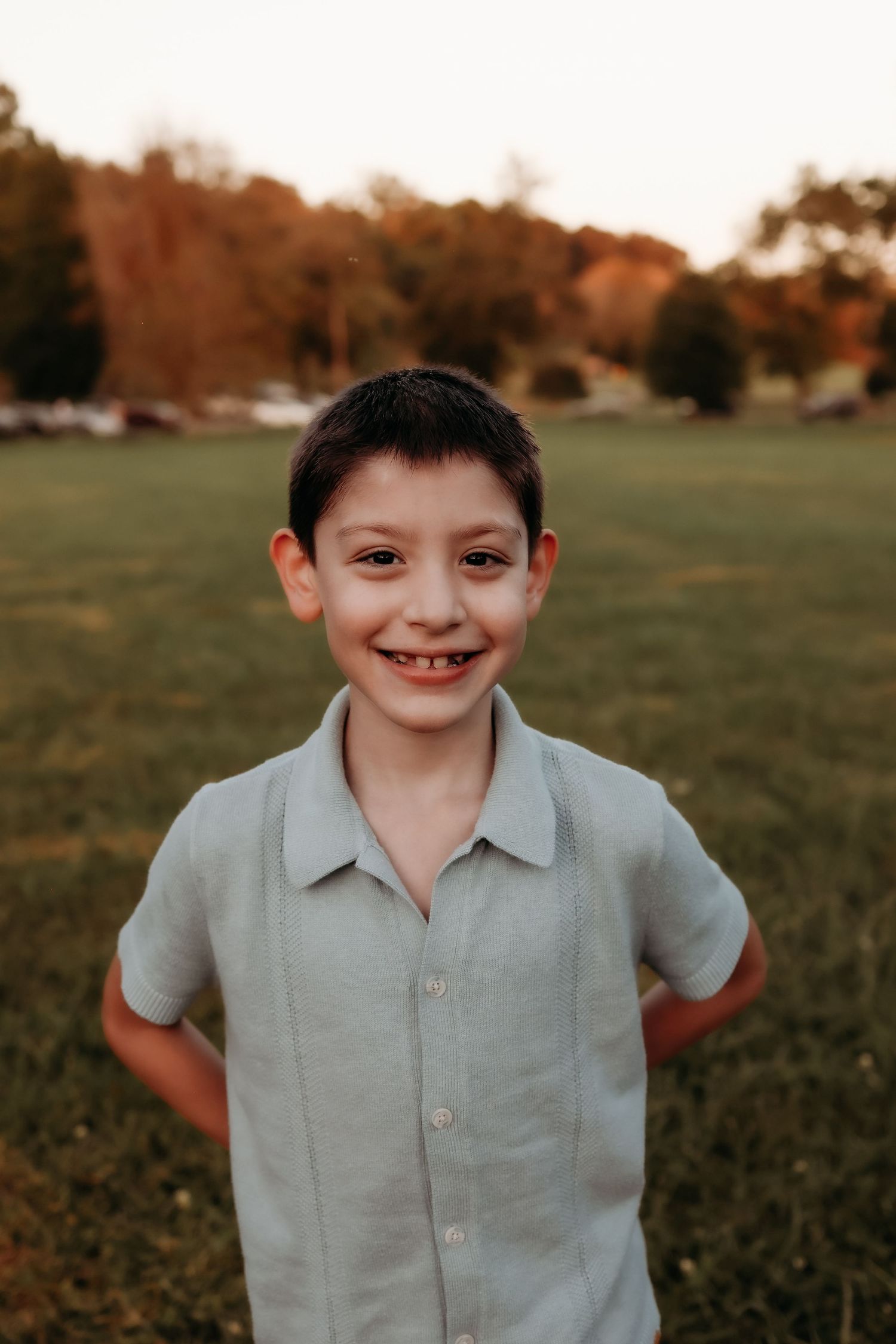 Smiling young boy in sage green shirt standing in green field at golden hour.