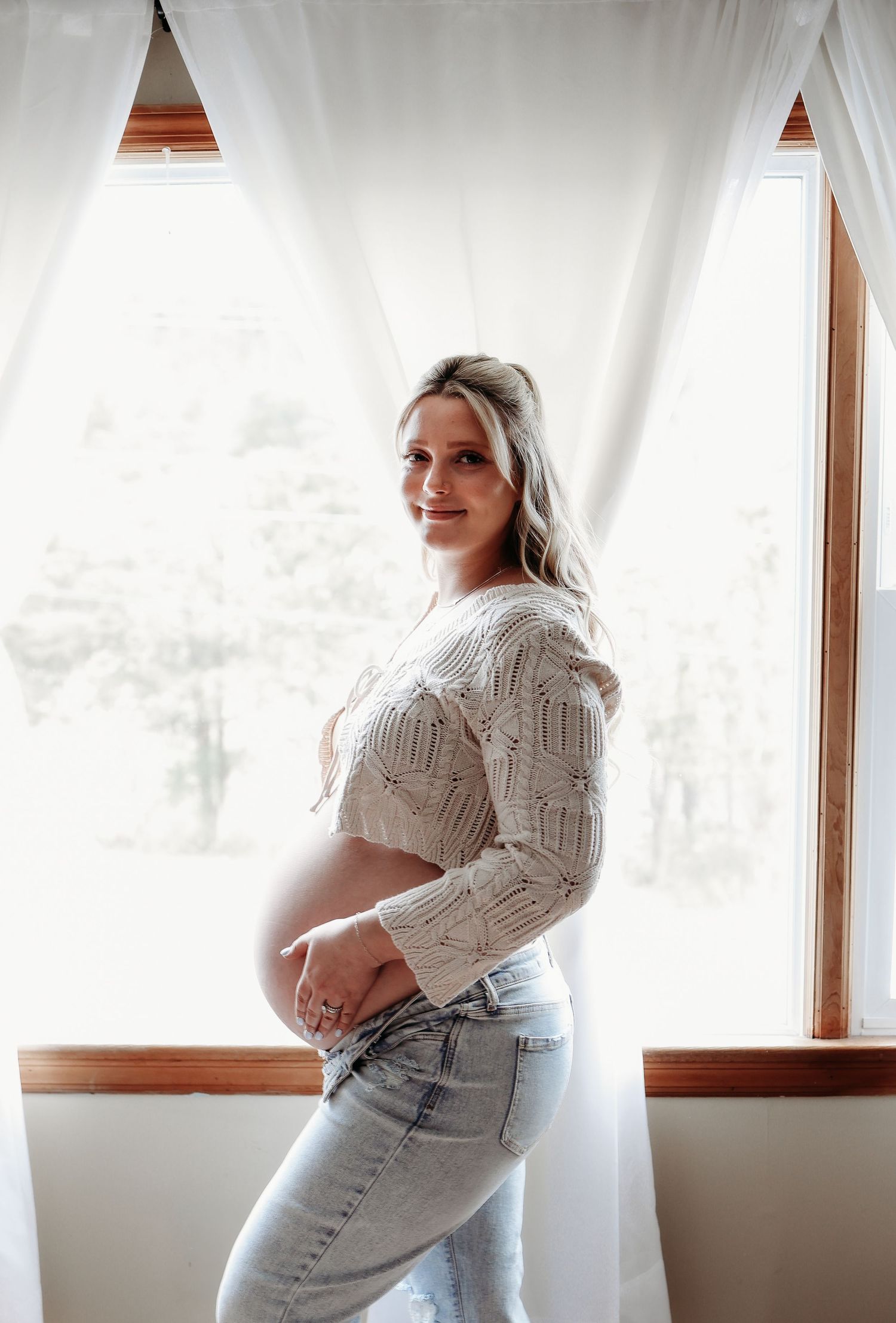 Pregnant woman in a crochet crop top and jeans posing sideways by a bright sunlit window.