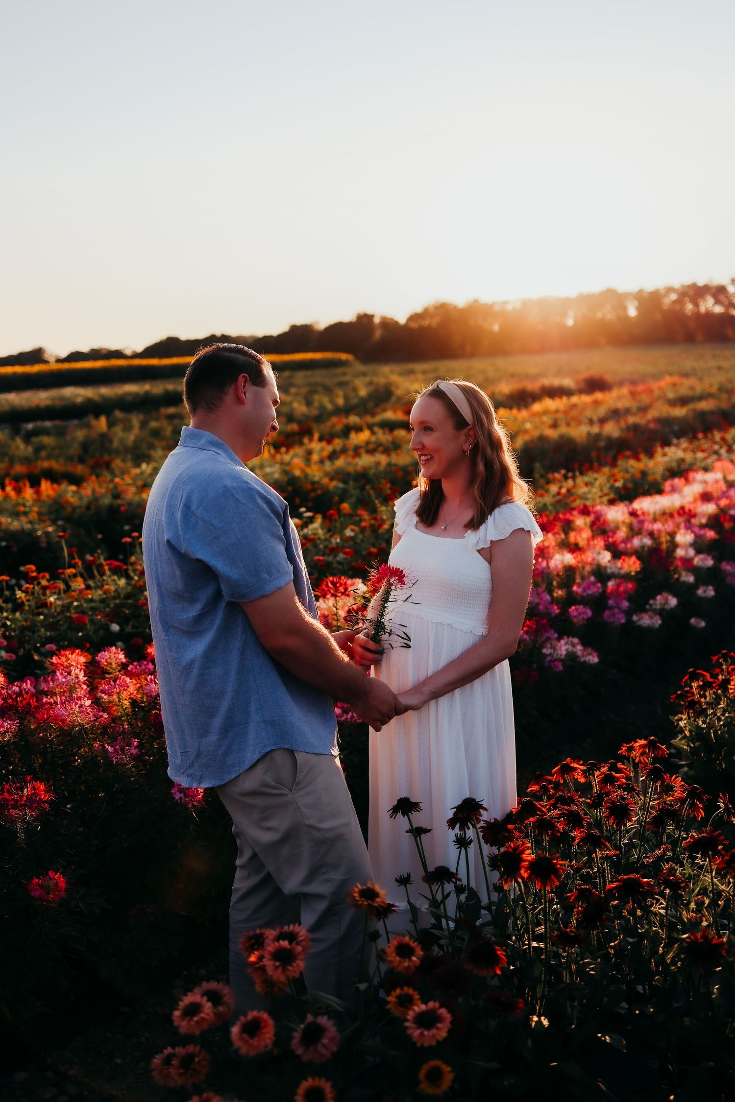 A couple shares a moment in flower field at sunset as warm golden light bathes the colorful landscape in Newton, New Jersey maternity shoot