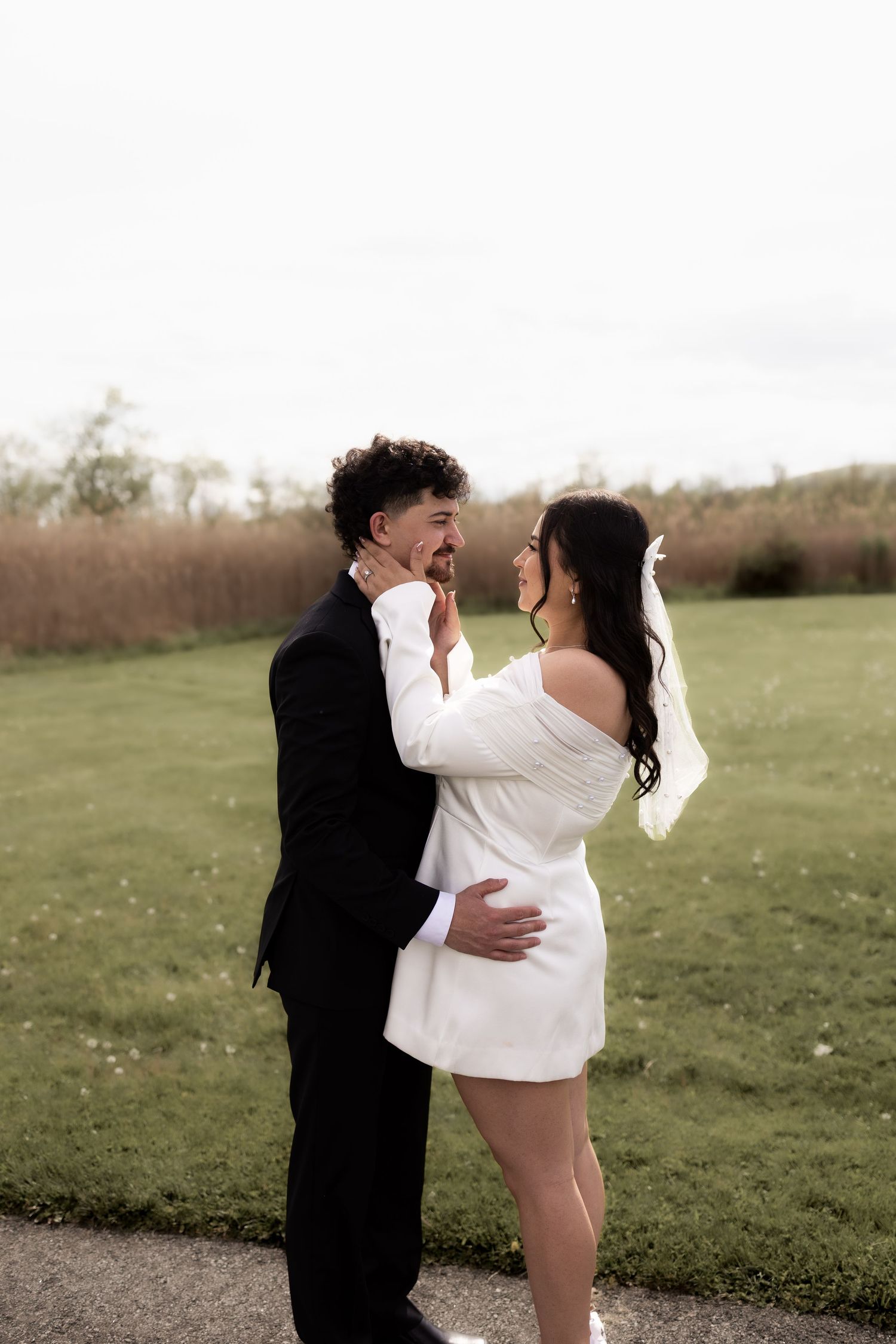 Bride in white mini dress and groom in black suit sharing a romantic moment outdoors on their wedding day.
