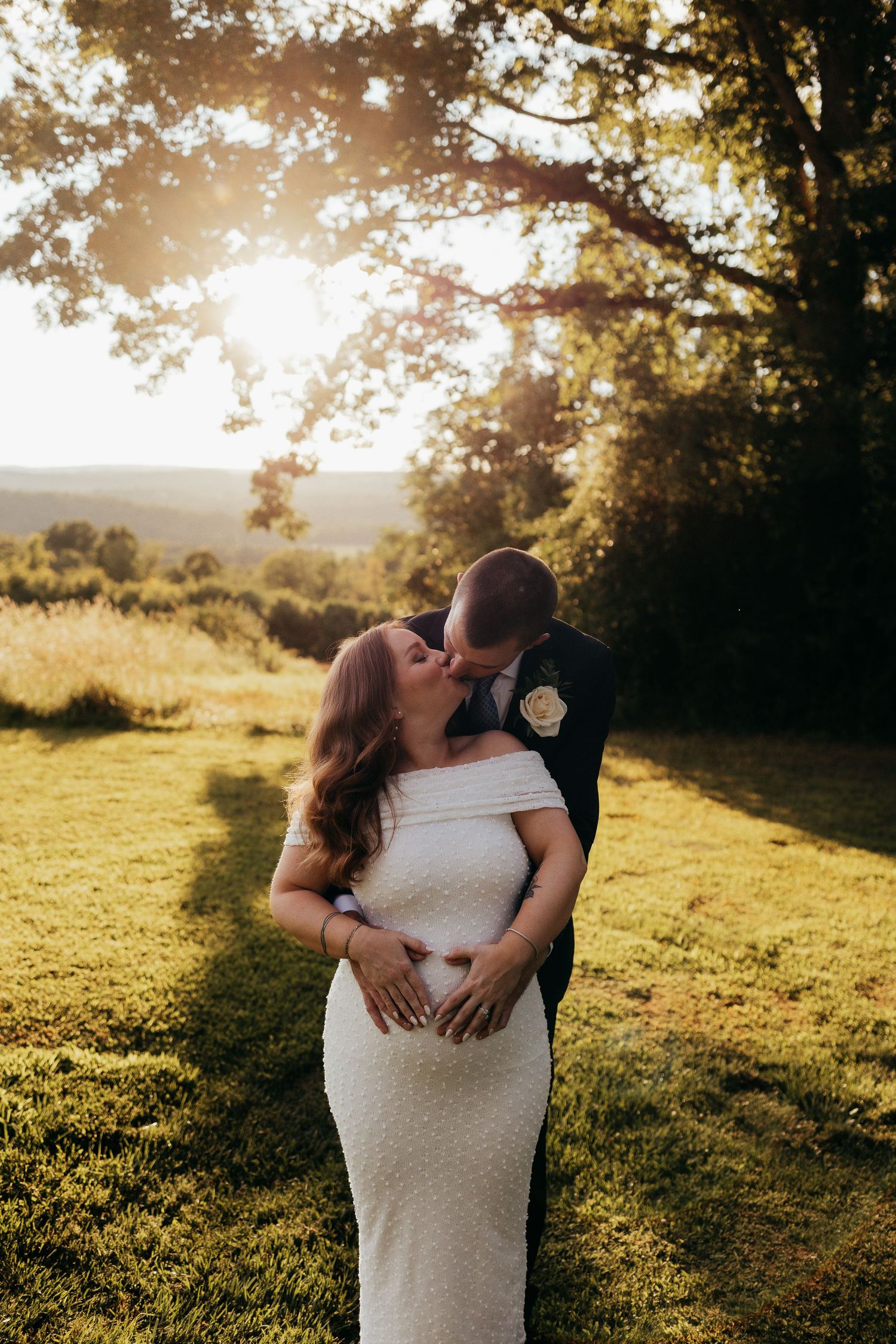 Pregnant bride in white off-shoulder gown kissing groom in suit at golden hour outdoors.