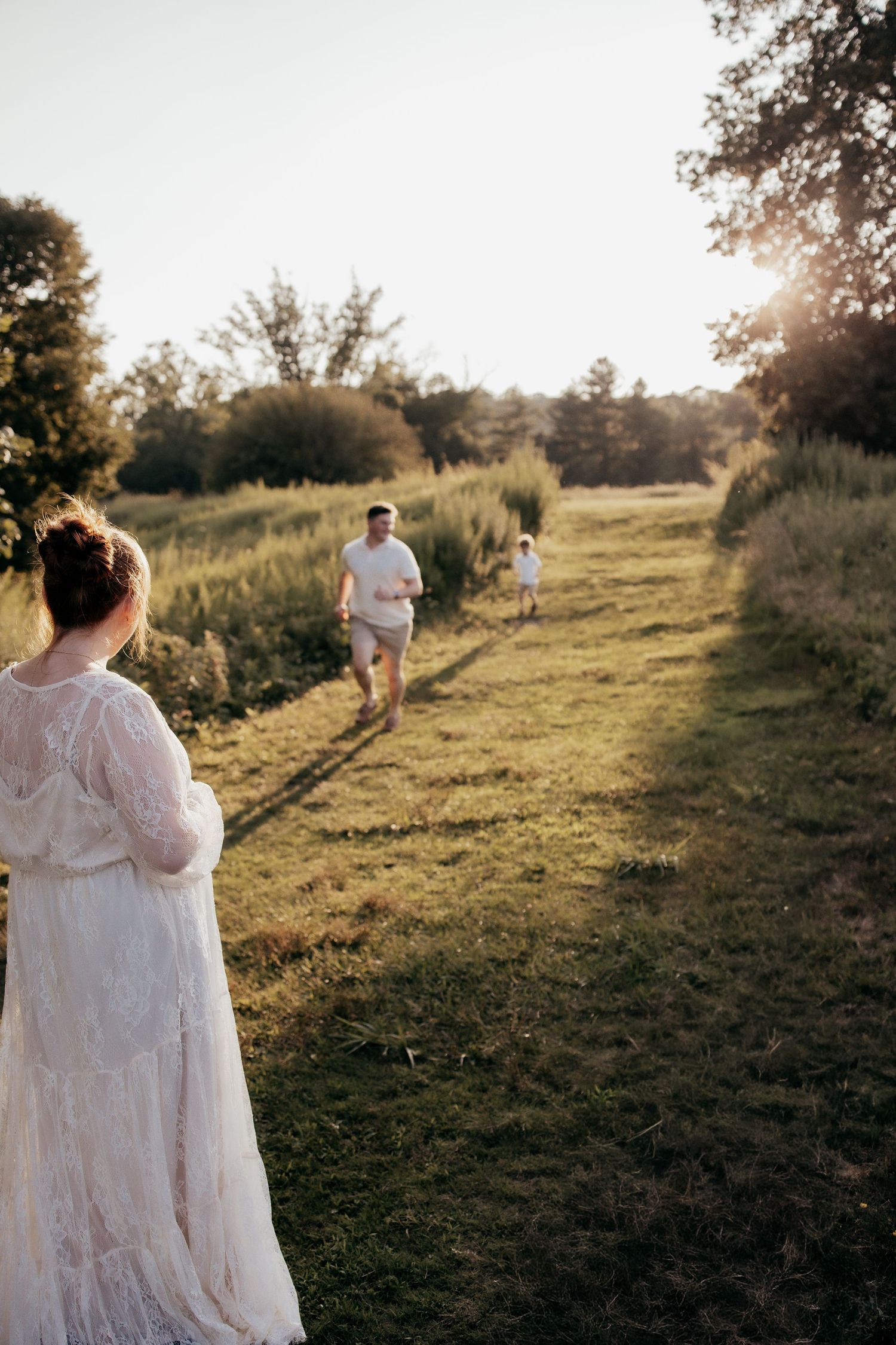 A pregnant woman in a white lace dress watches her family run toward her through a sunlit green field at golden hour.