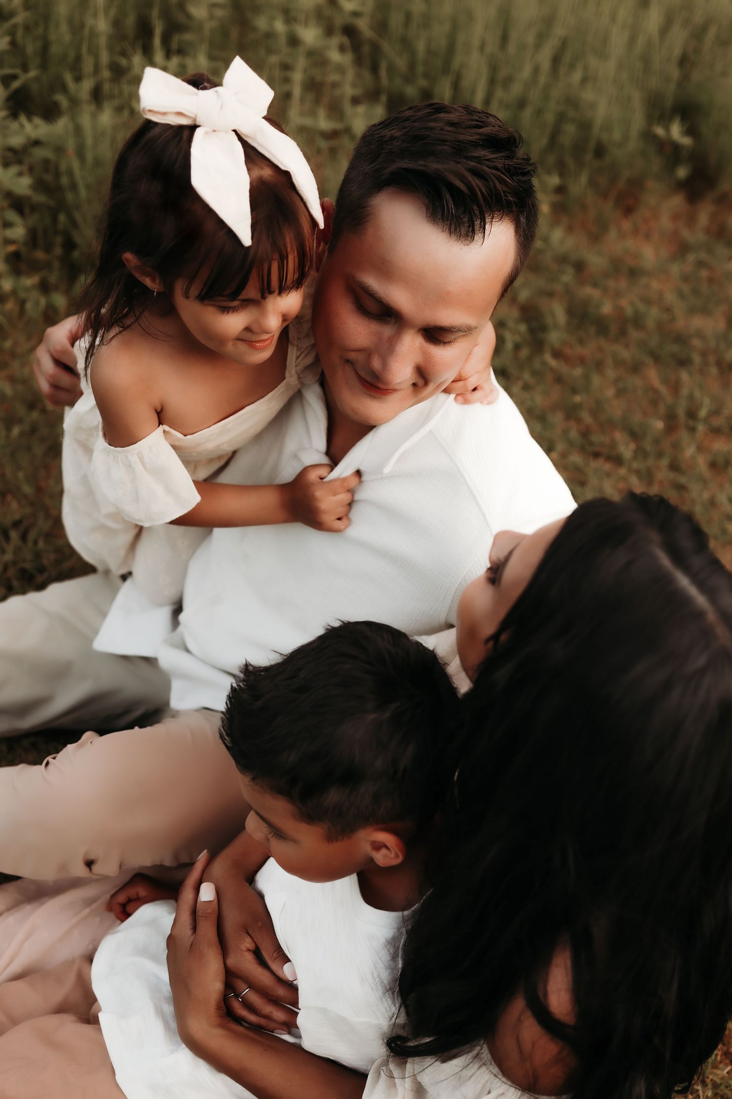 Happy family of four in white outfits sitting together in a green field, sharing a joyful embrace.