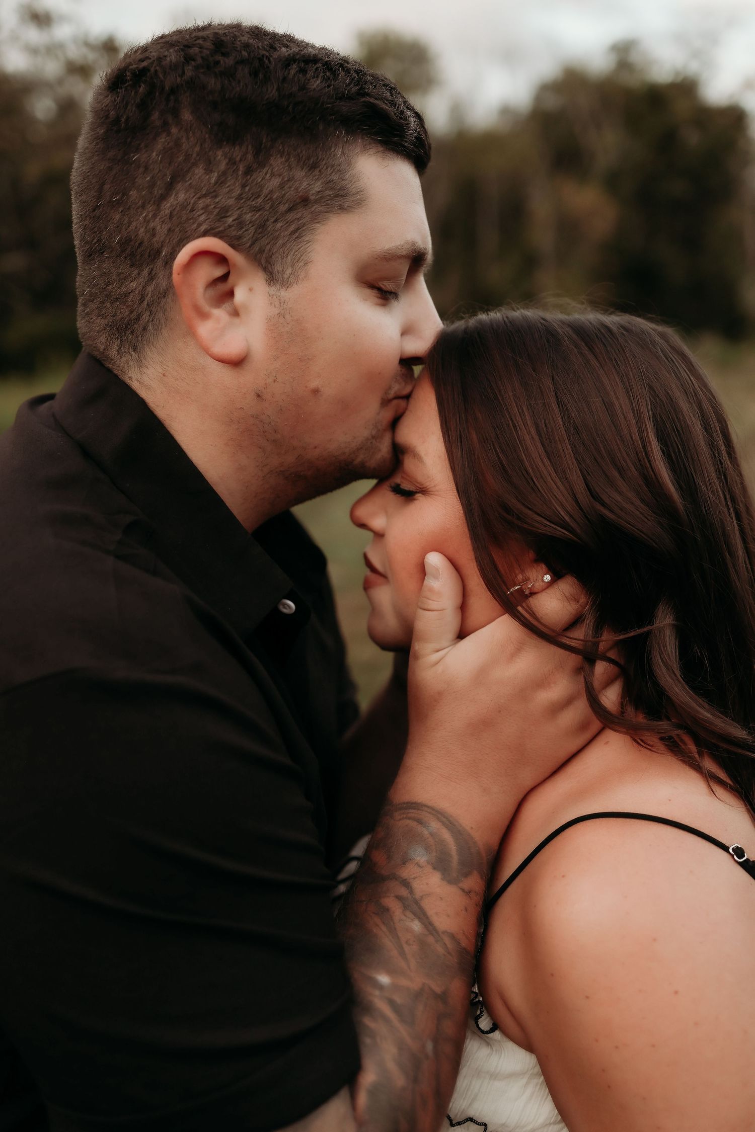 Man with tattoos kissing woman on forehead tenderly during an outdoor couples portrait session at dusk.
