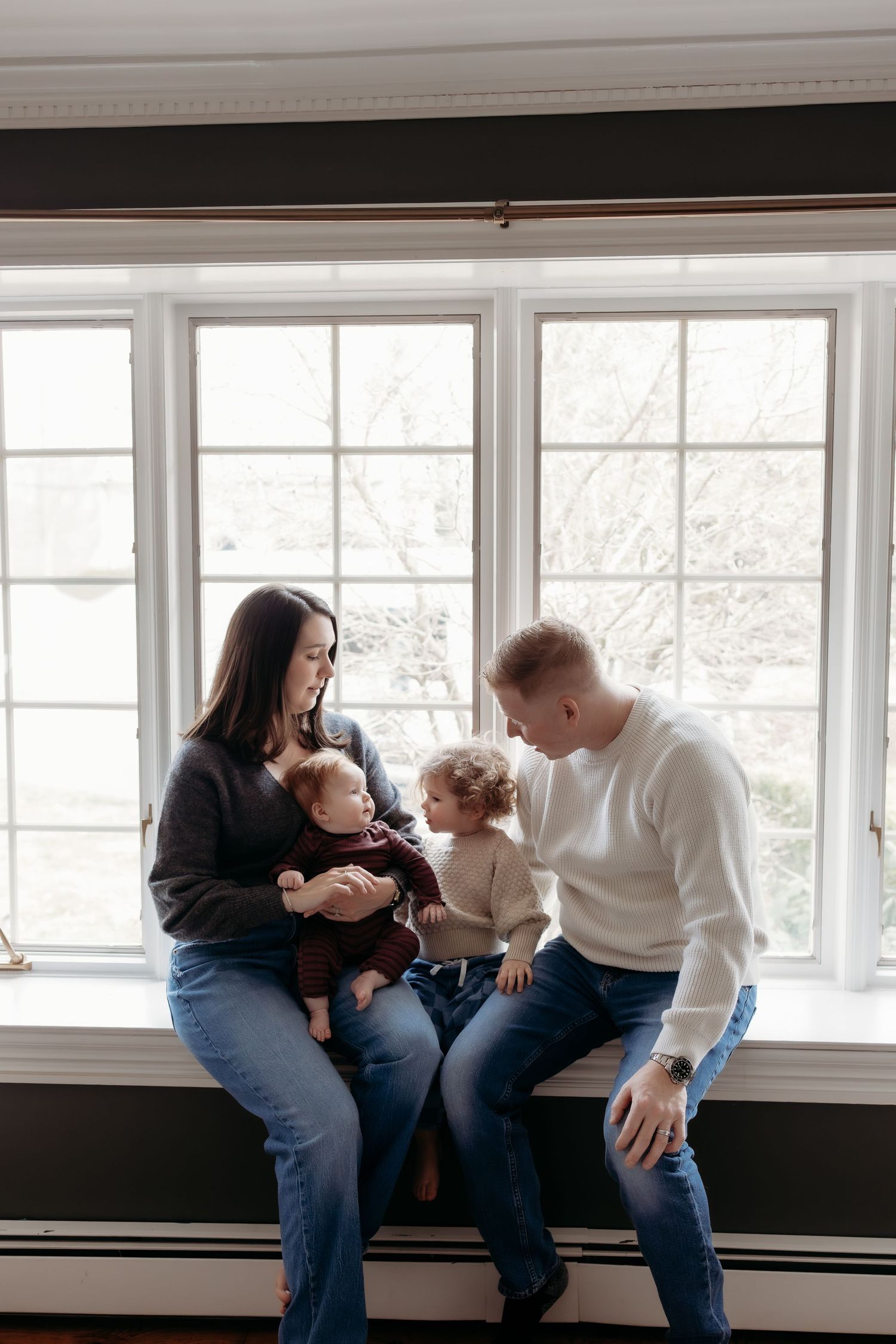 Family of four sitting on a window sill, parents holding two young children in a bright, cozy home setting.