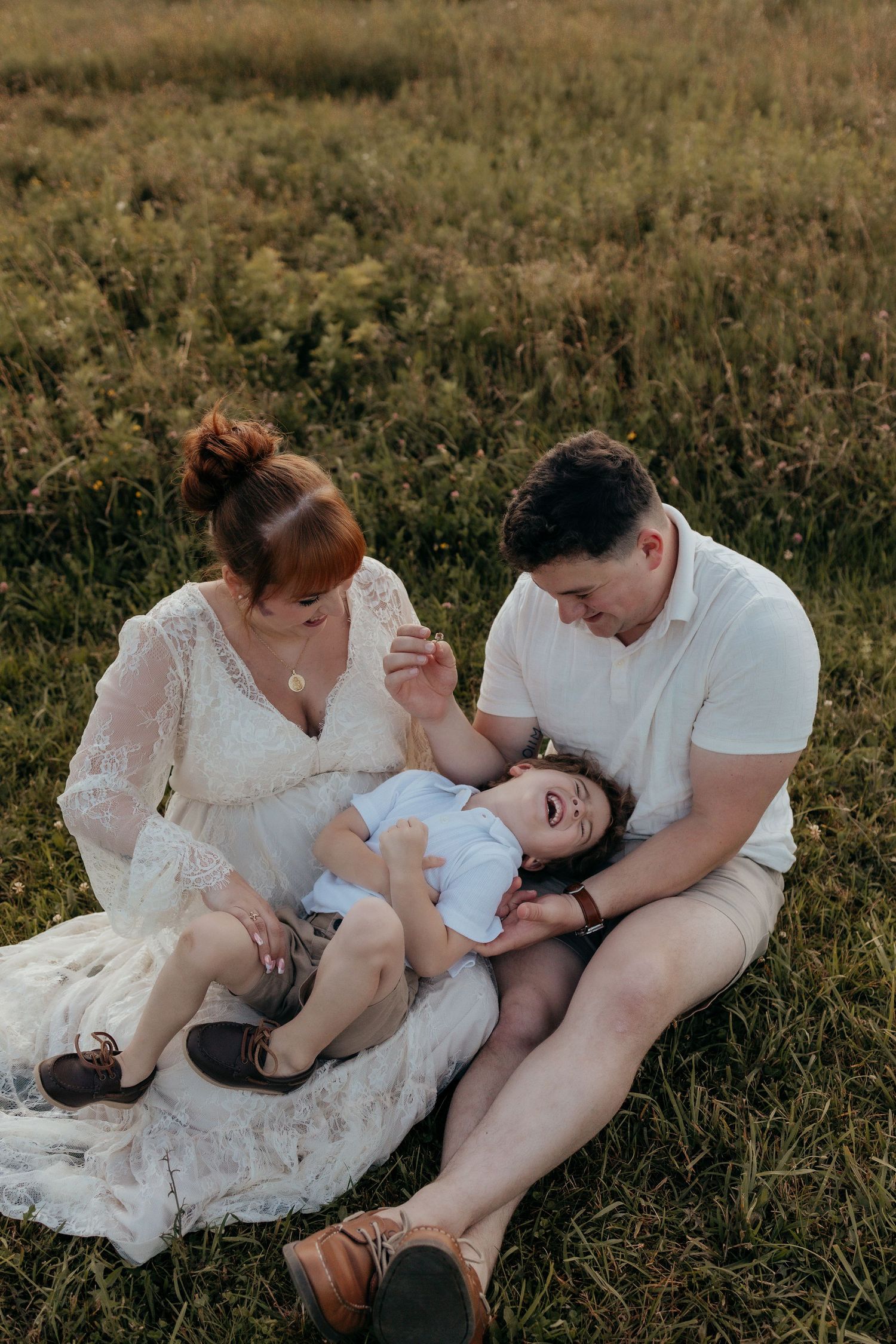 A family of three sits together in a grassy field, parents smiling at their laughing toddler.