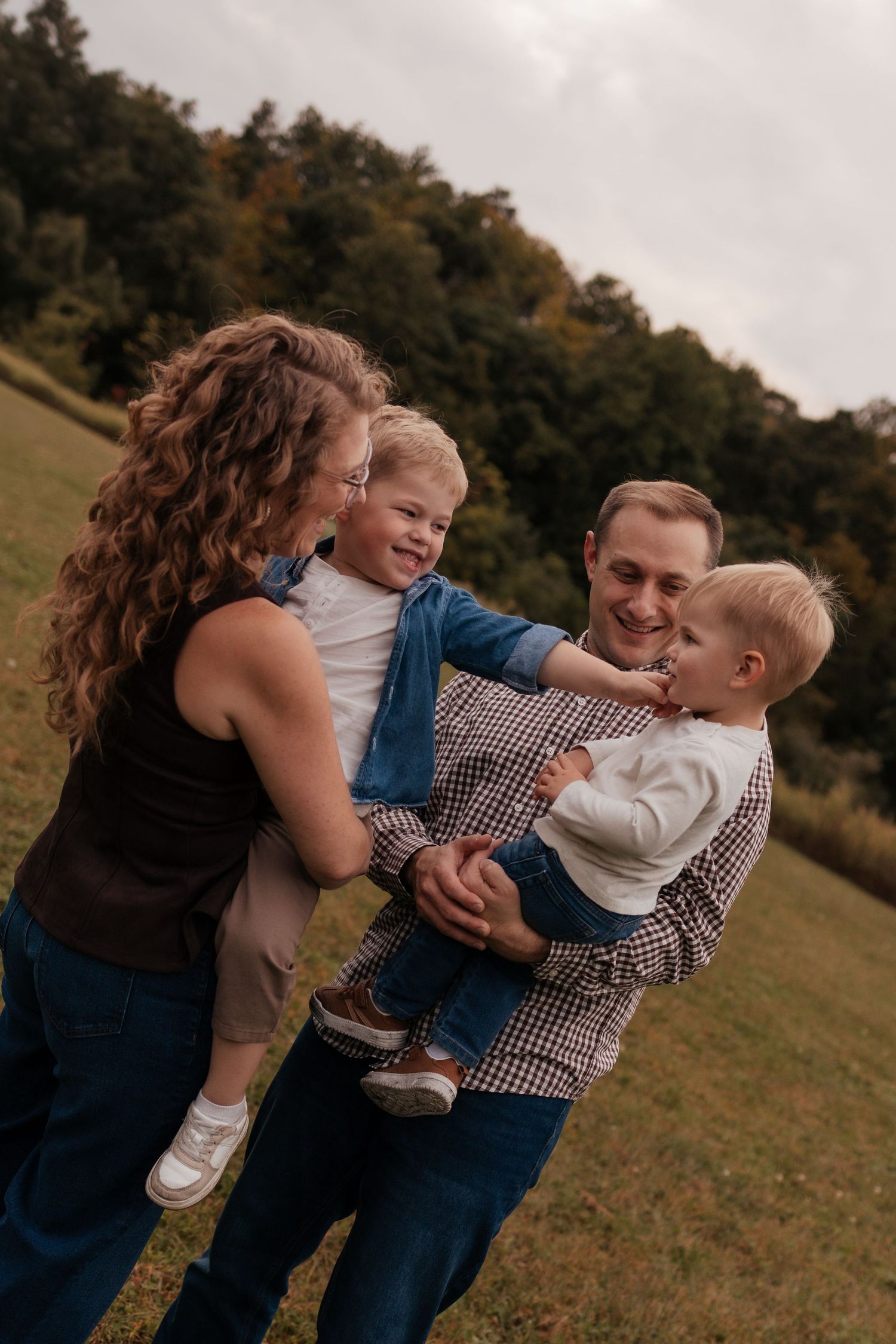 A happy family of four enjoying outdoor time, with parents holding two young toddler boys in a green park setting.