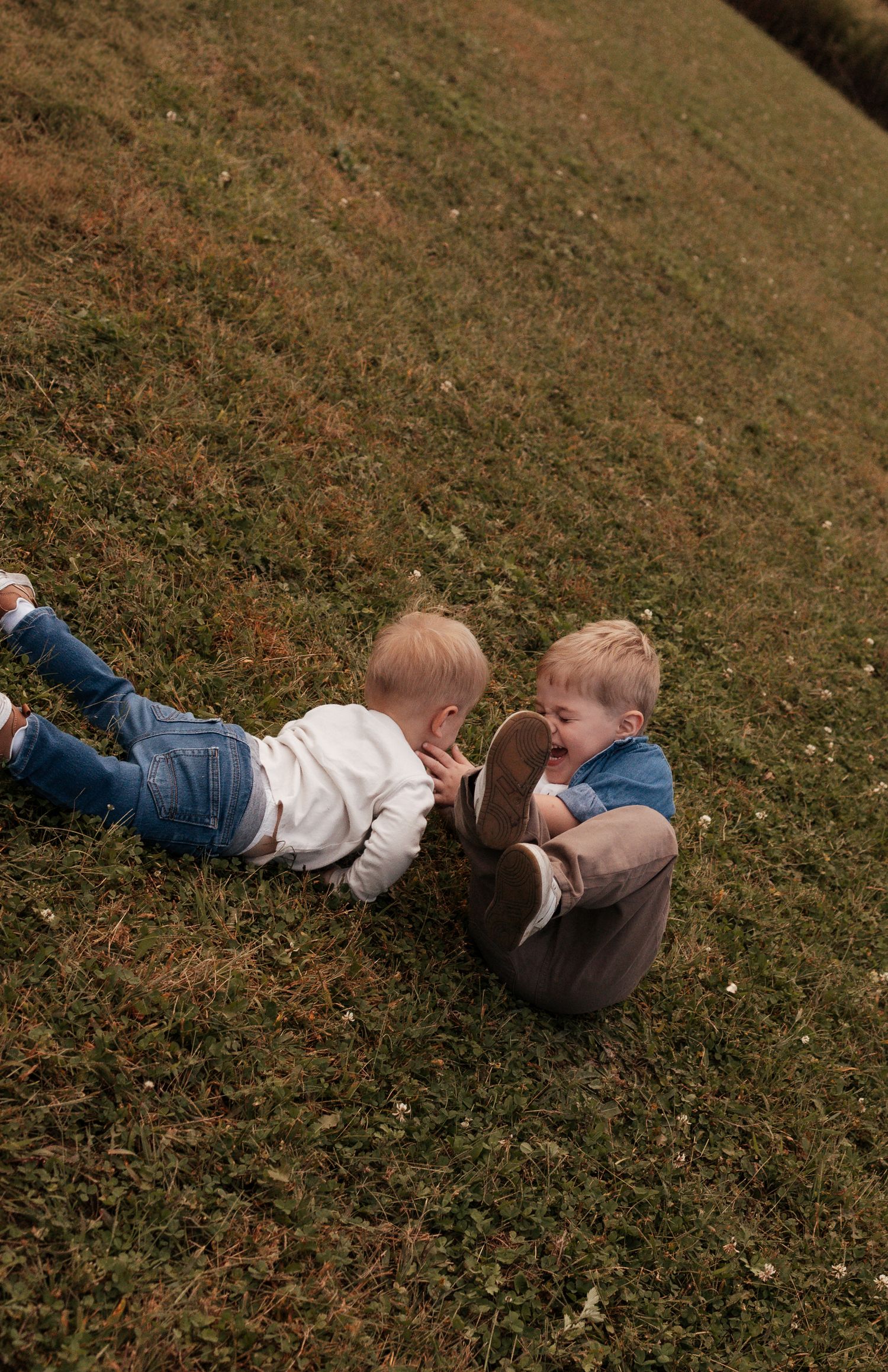 Two young blonde toddlers playing together on green grass, one lying down and one sitting up, sharing a joyful moment outdoors.