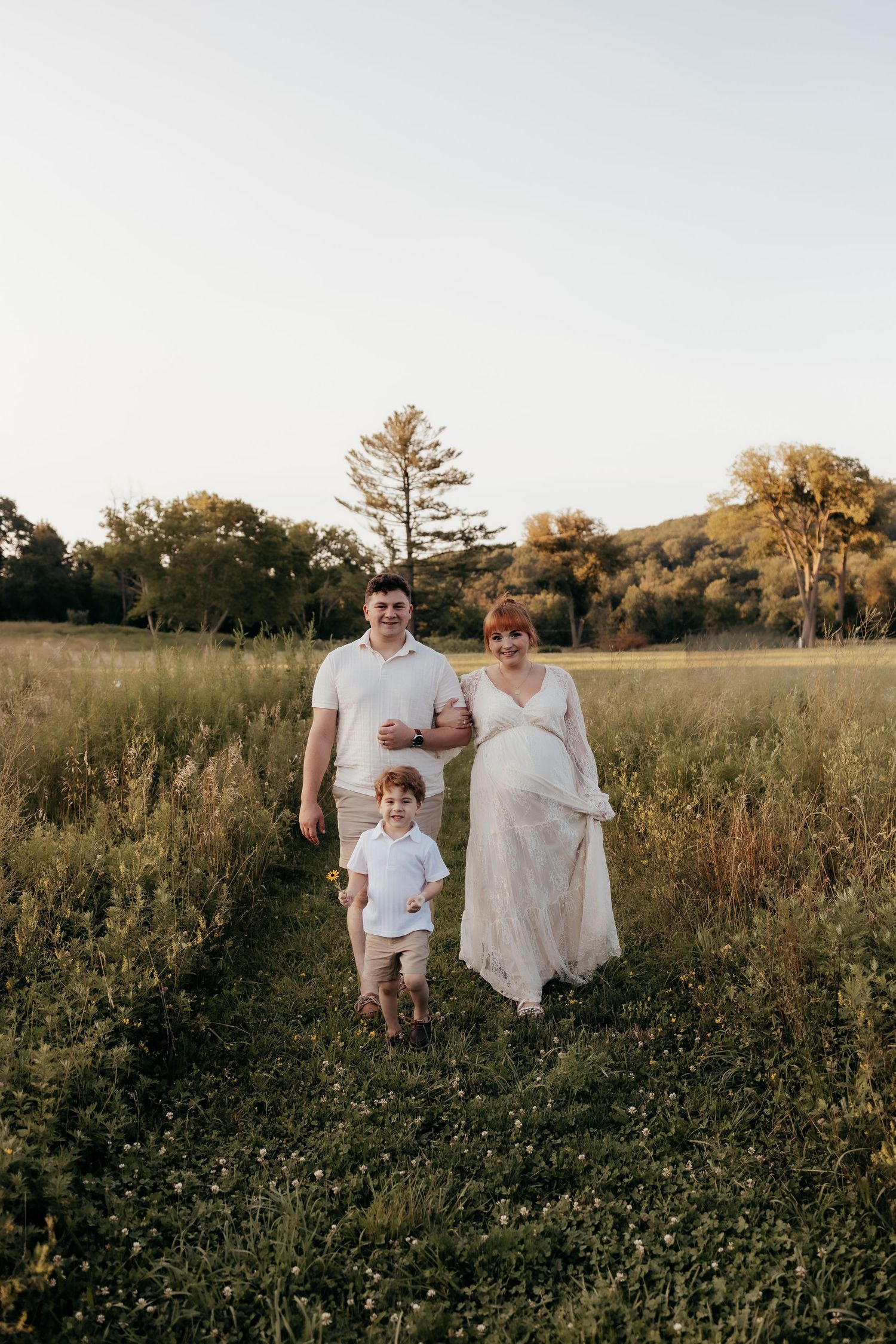 Pregnant woman in lace gown walks with husband and toddler son through sunlit green meadow path at golden hour.