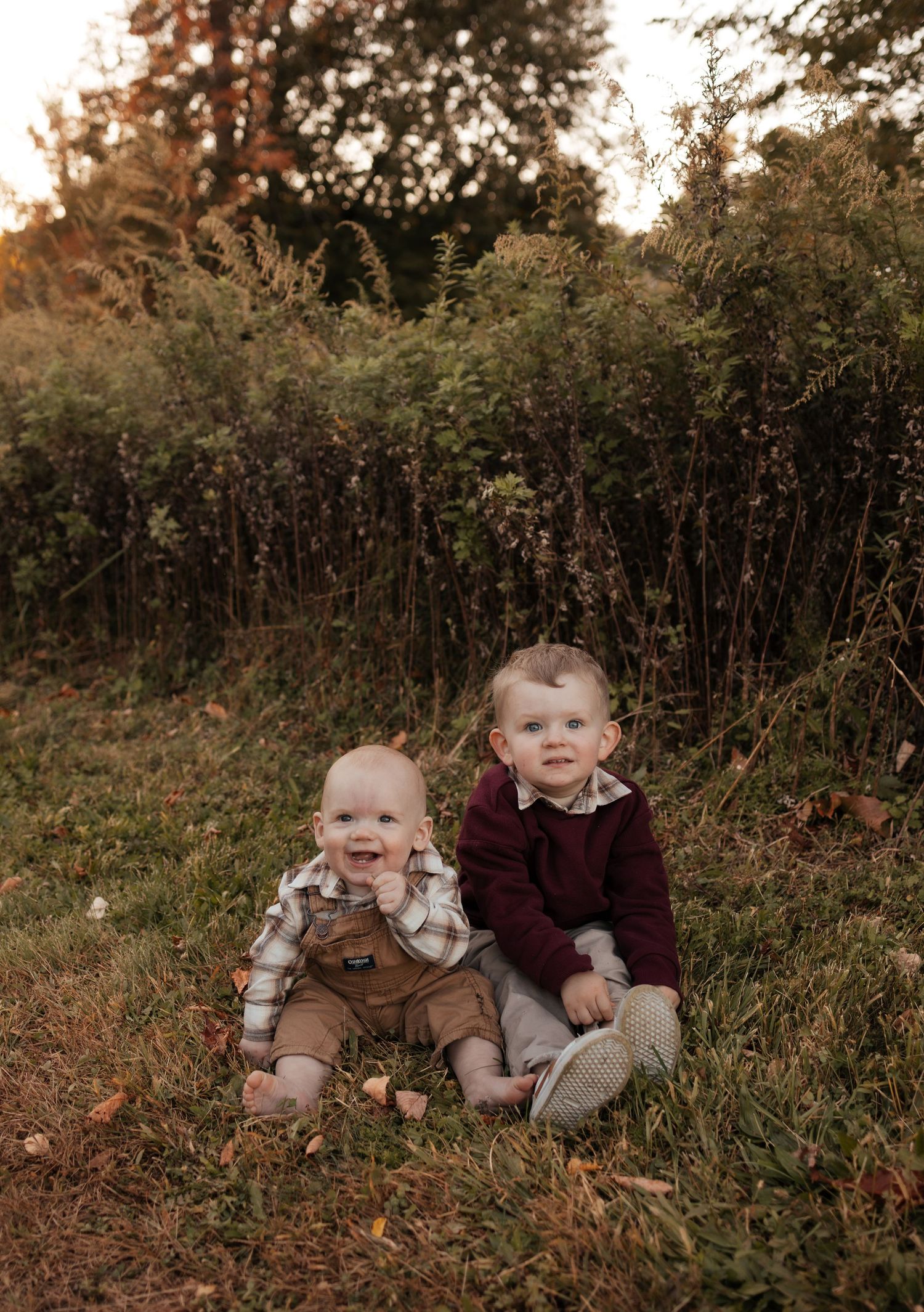 Two smiling toddlers in fall outfits sit together on a grassy lawn dotted with leaves in front of golden autumn foliage.