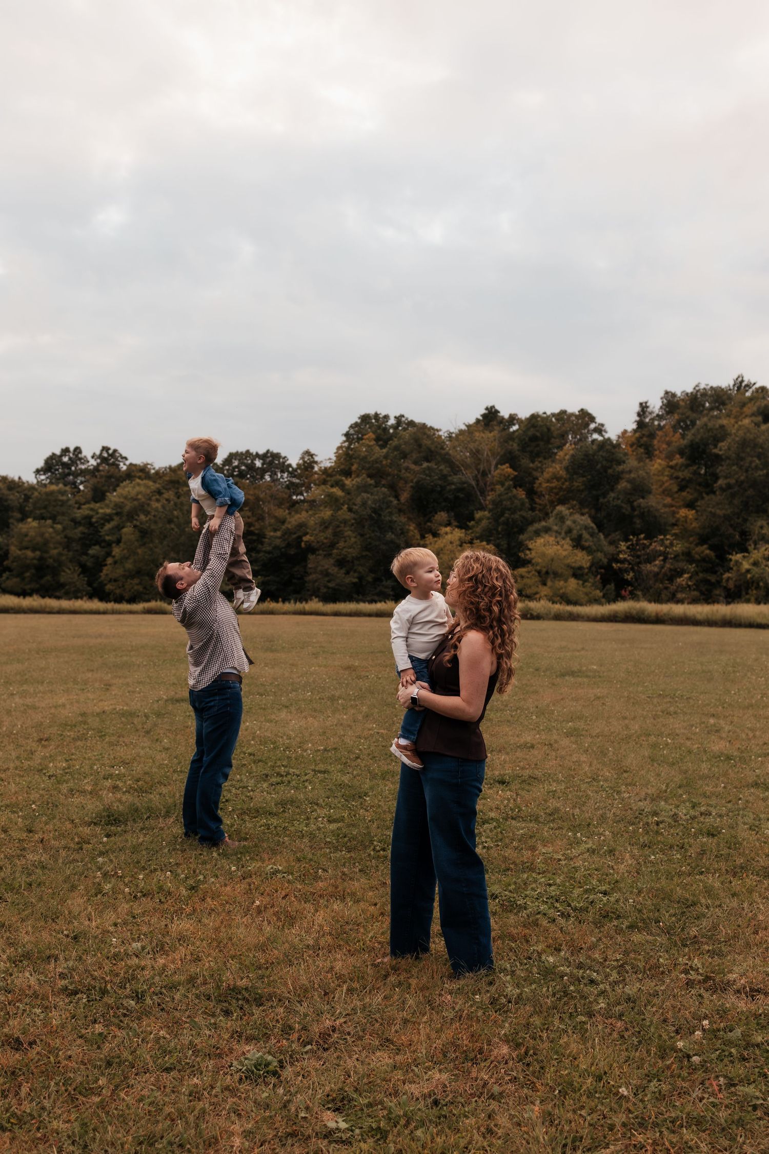 Family of four enjoying a fall day in an open field, with dad lifting child overhead and mom holding toddler.