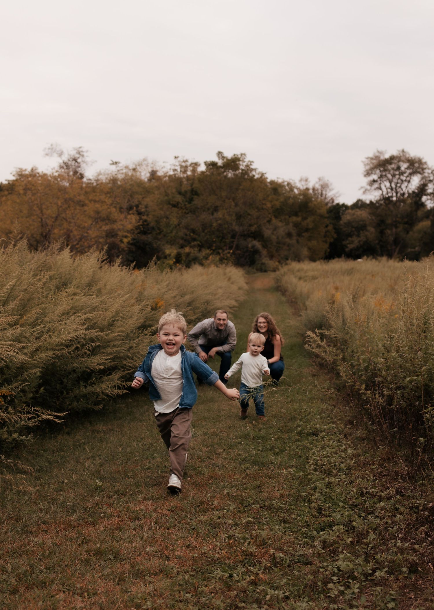 Two young children run joyfully down a grassy path while their parents watch and smile in an autumn field.