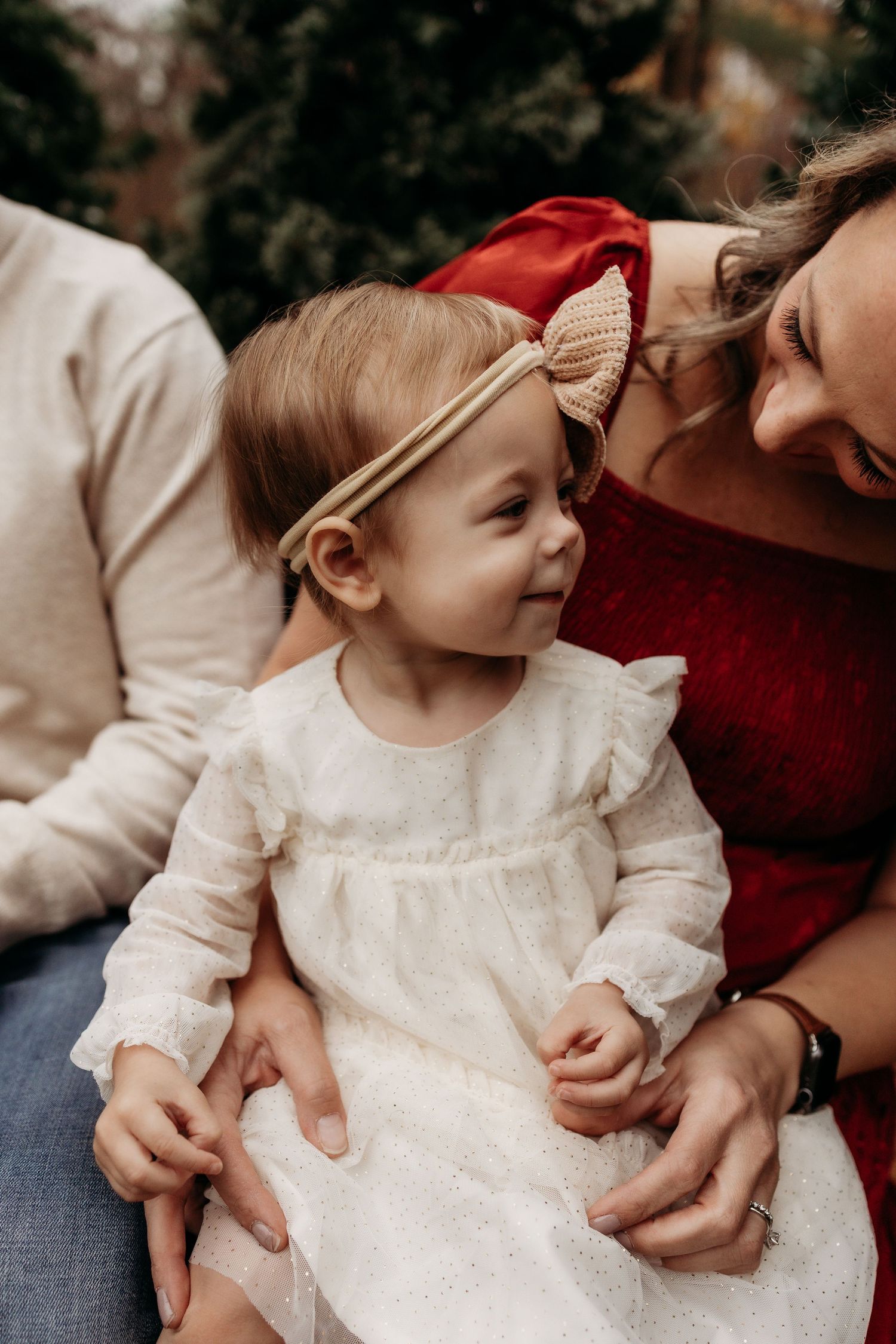 A baby girl in a white lace dress with a bow headband sits on her mothers lap outdoors near evergreen trees.