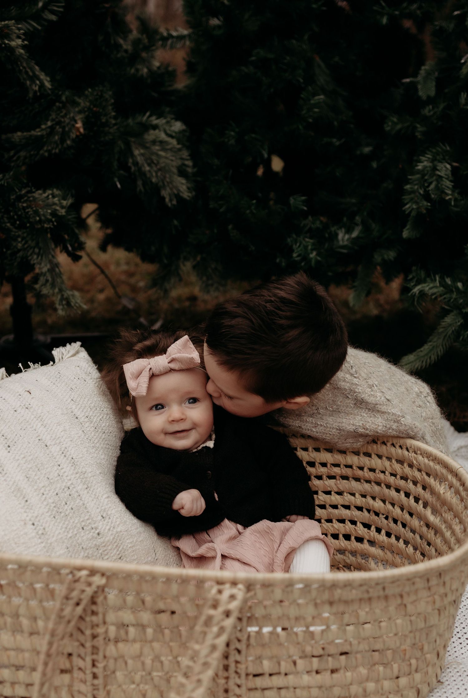 Older child kissing baby sibling in a woven basket, baby wearing pink bow headband, surrounded by evergreen trees.
