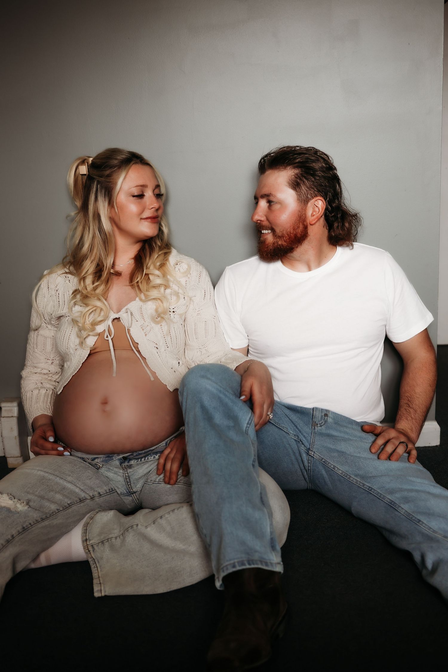Expecting couple in casual white tops and jeans sitting together, woman showing pregnant belly, smiling at each other.