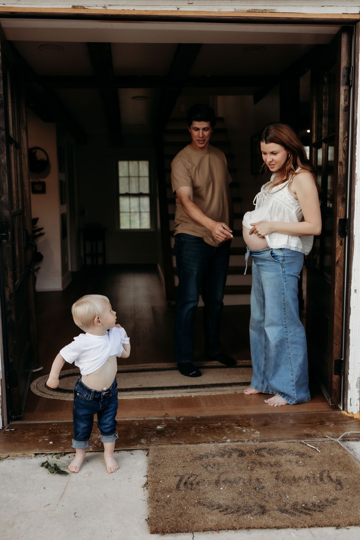 Young family with toddler standing at farmhouse doorway, pregnant mother cradling belly beside partner.