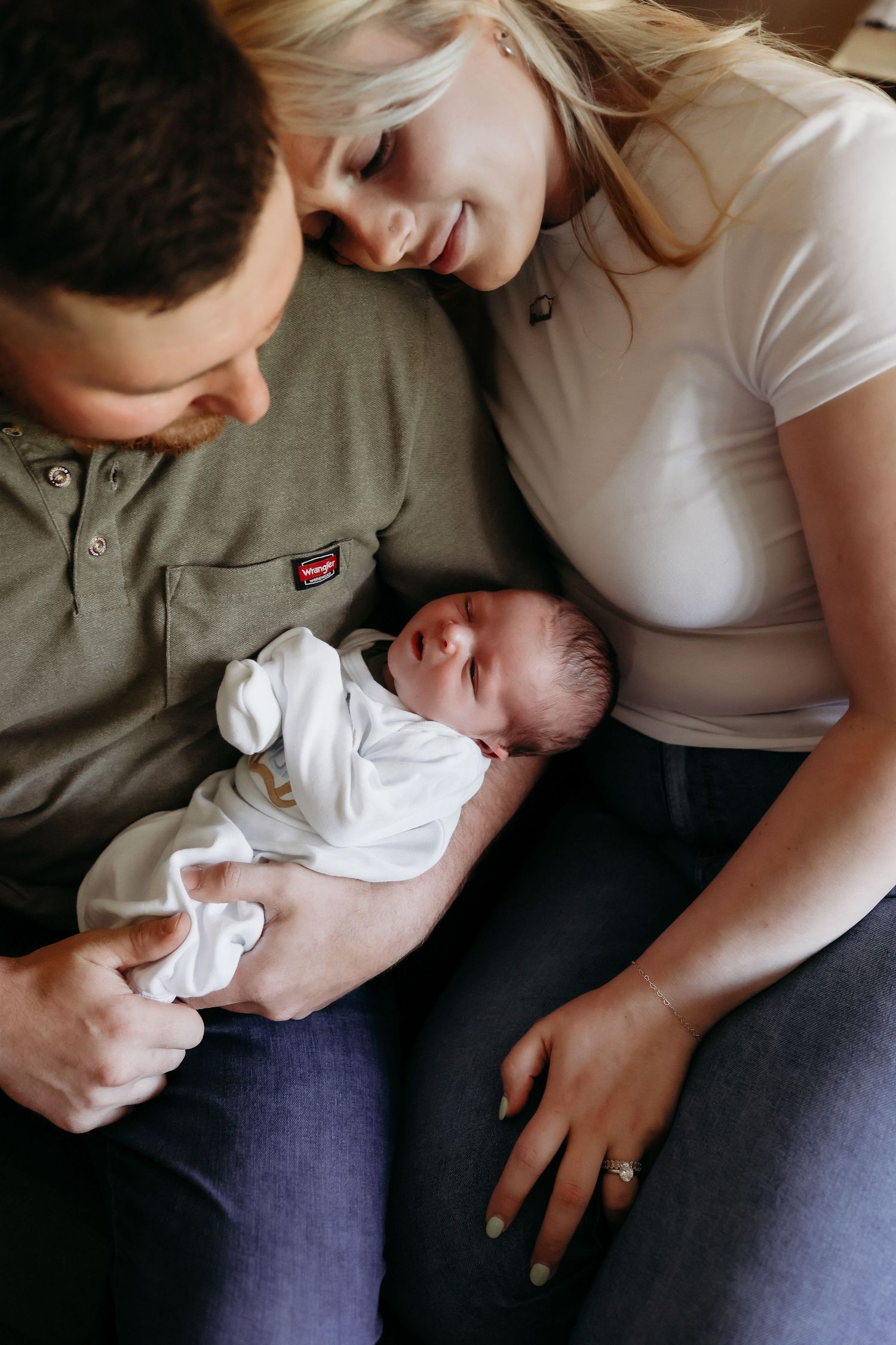 Smiling parents in olive and white tops lovingly gazing down at their sleeping swaddled newborn.