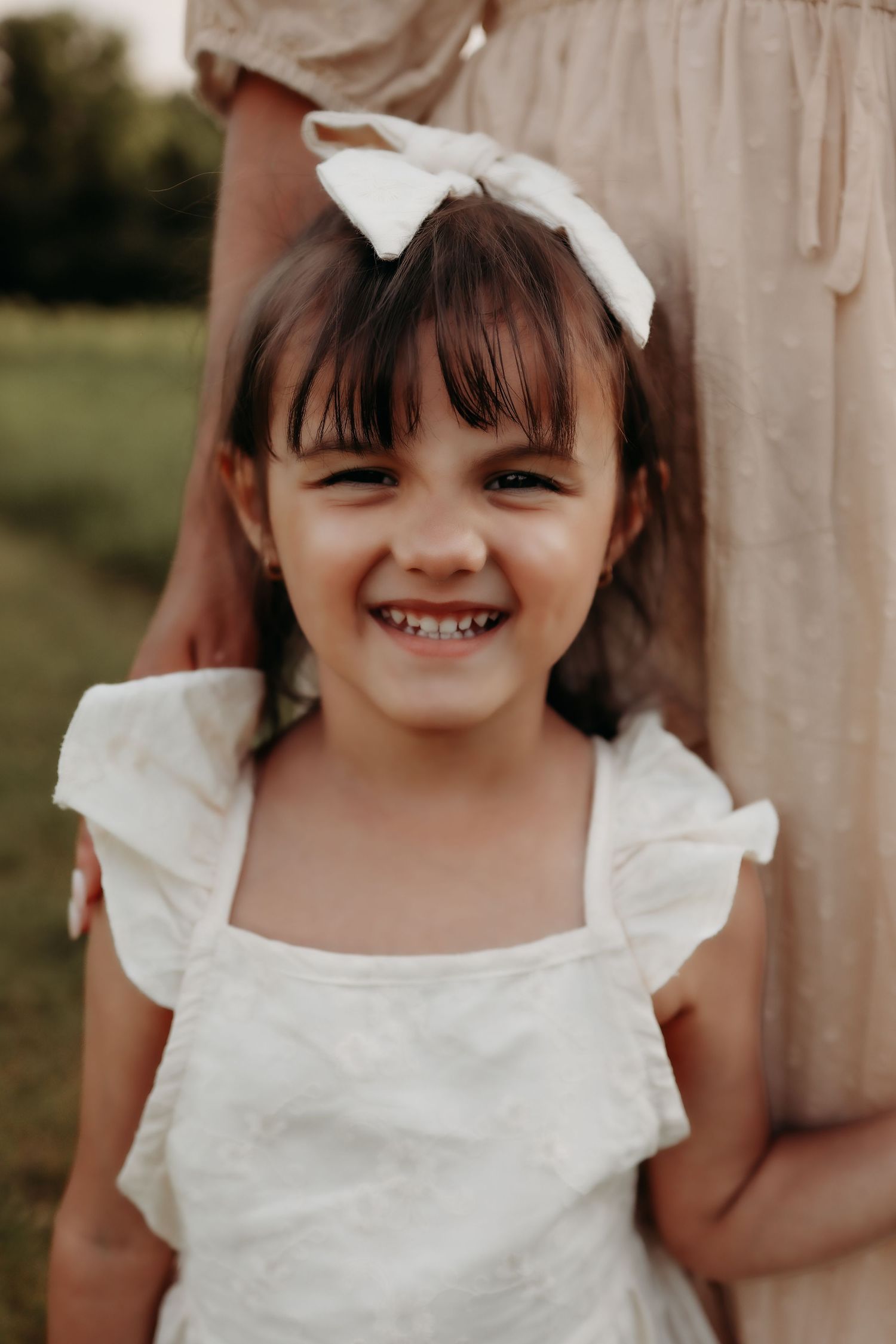 Smiling young girl with dark bangs wearing white dress and bow hairclip outdoors.
