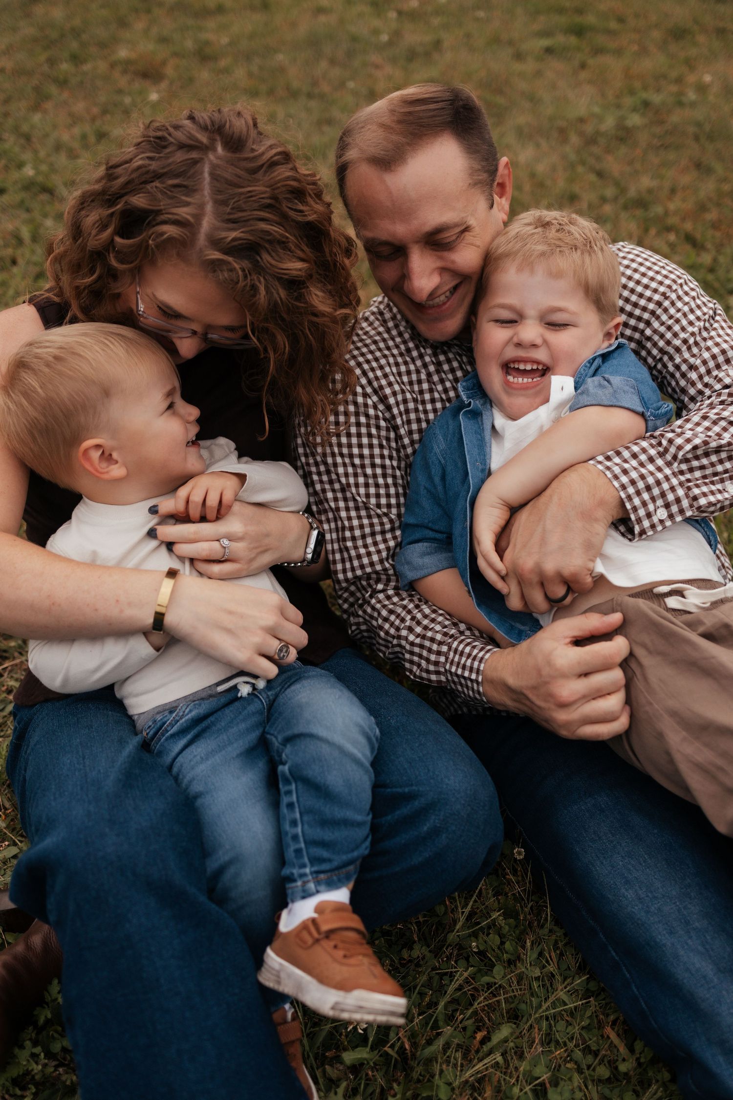 A joyful family of four sits on grass, parents cuddling and laughing with two young boys in casual denim outfits.